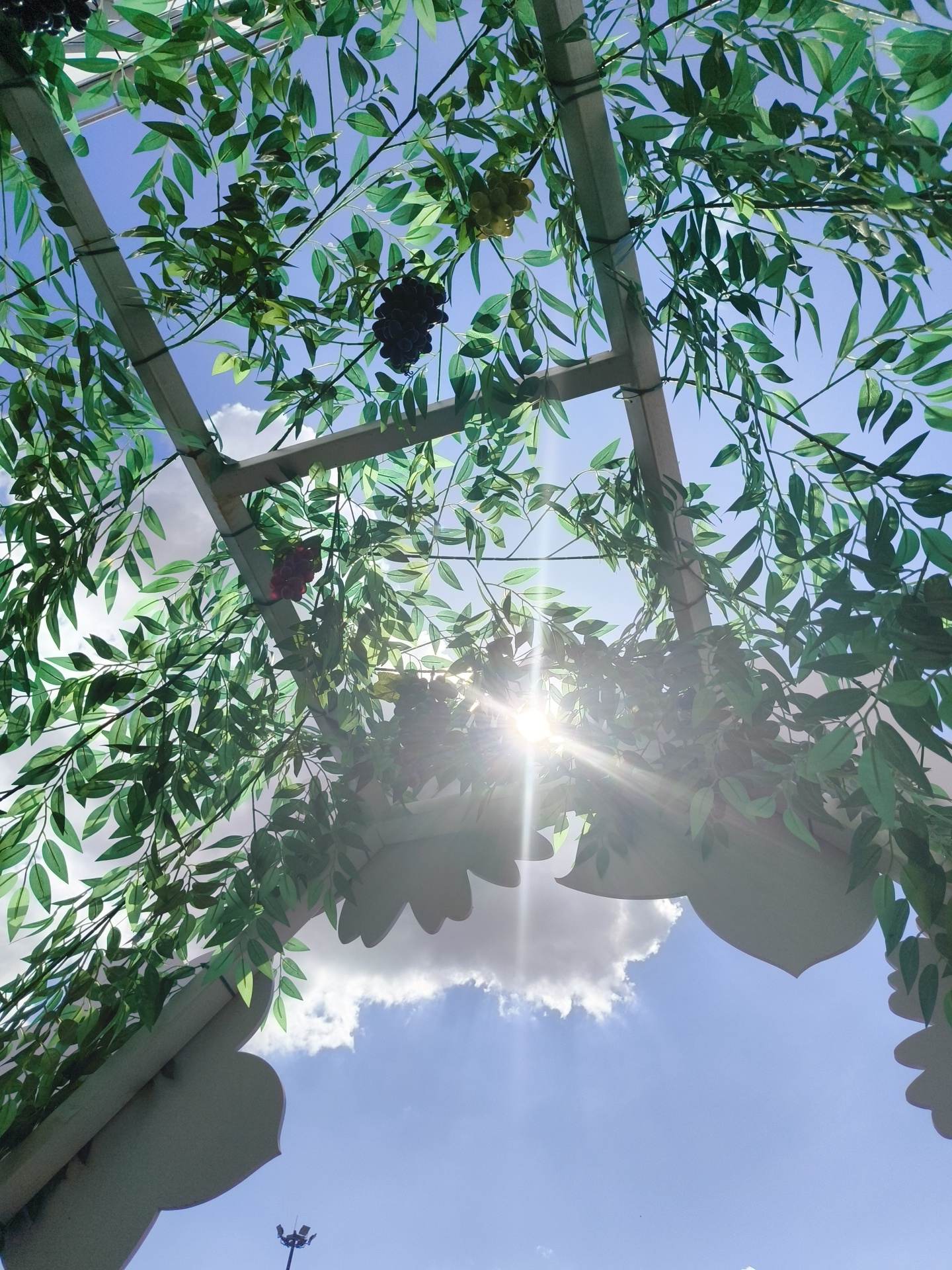 This is a low-angle, full shot of a structure with artificial greenery and grapes hanging from it, set against a blue sky with clouds. The sun is visible through the greenery, creating lens flare. The structure appears to be part of an outdoor space, possibly a garden or an event venue. In the distance, a tall pole with lights is visible against the sky. The context of Desnogorsk, Russia, is not visually discernible from the image itself.