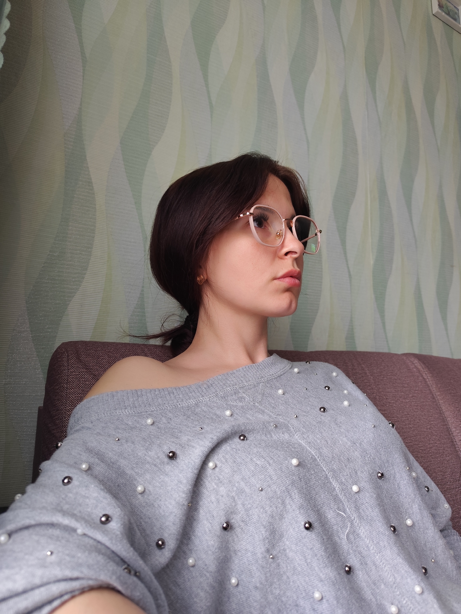 A young woman with dark hair is sitting on a brown couch in front of a wall with green and white patterned wallpaper. She is wearing glasses with pink frames and a light gray sweater adorned with both silver and white pearls. The sweater is partially off her left shoulder. Her hair is pulled back into a low ponytail, and she is wearing small gold earrings. She is looking to her right with a neutral expression. The location, Фоки, Russia, cannot be determined from the provided image.