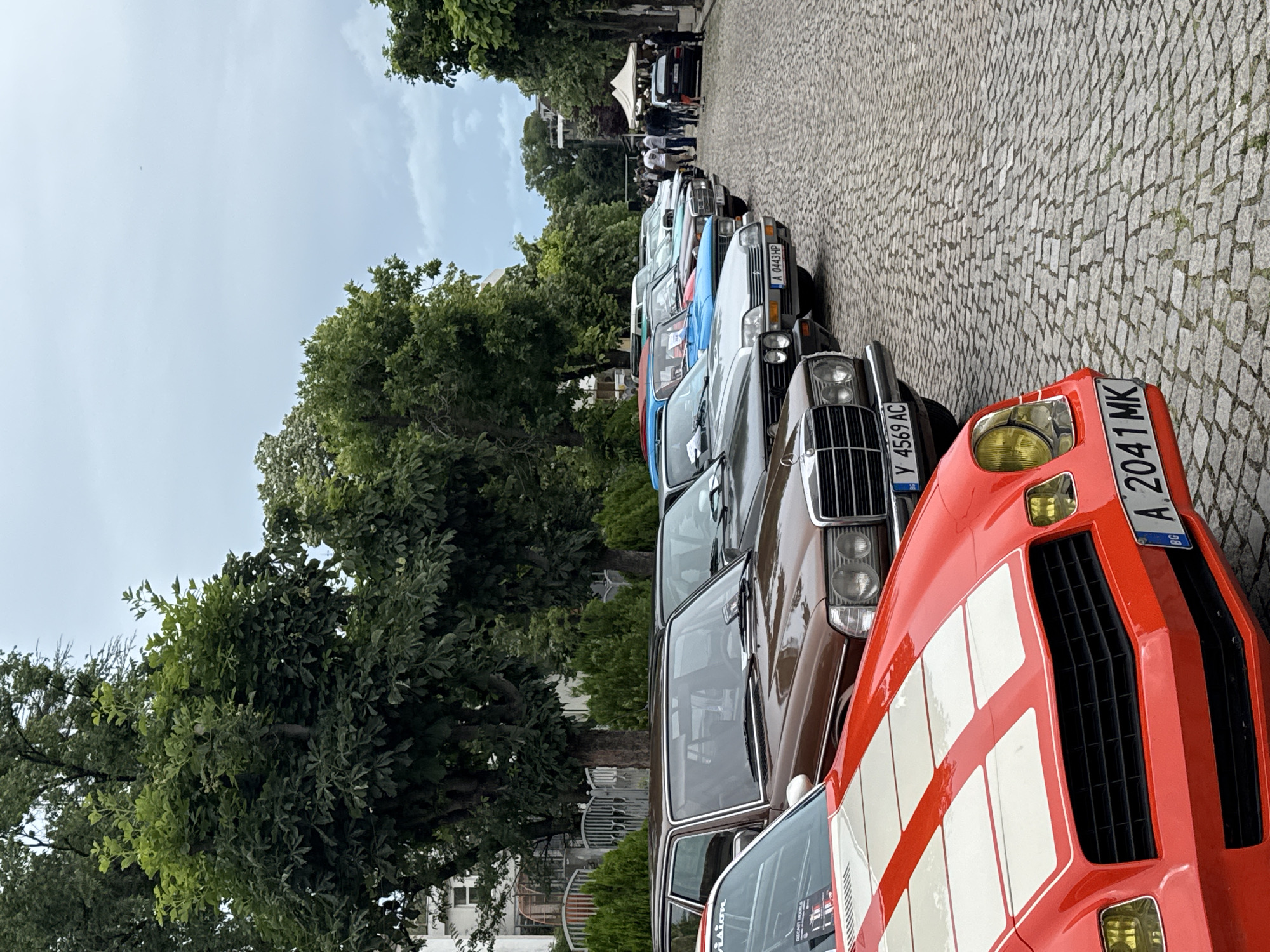 In Tsarevo, Bulgaria, a street lined with cobblestones and trees displays a collection of vintage automobiles. The cars, parked in a row, include a prominent red Chevrolet Camaro with white racing stripes, a brown Mercedes-Benz, a gray Citroen, and several other classic models. A white tent is visible in the background, suggesting an event or gathering. Several people are standing around, observing the vehicles. The sky is overcast, and the overall atmosphere appears to be that of a car show or exhibition.