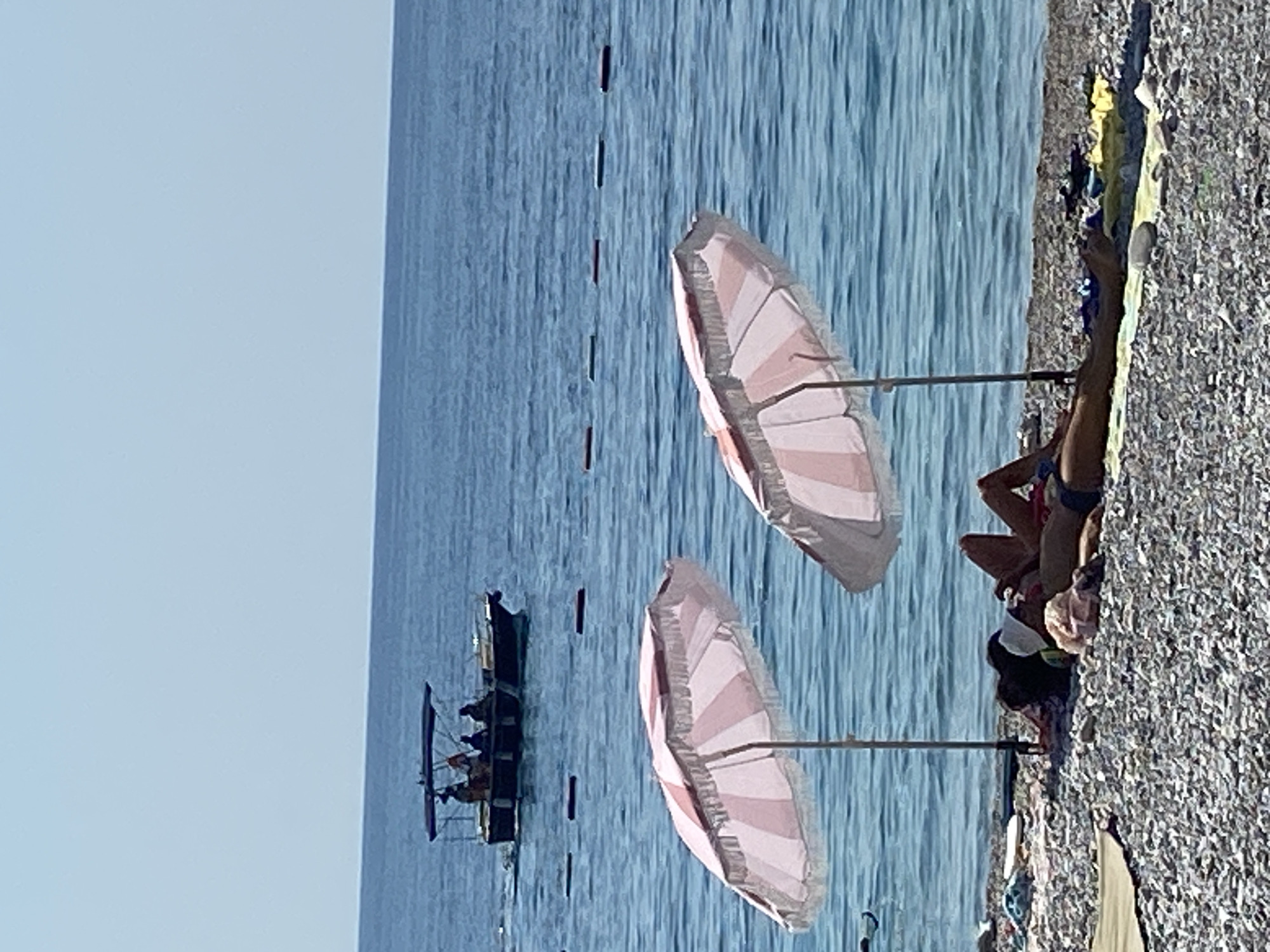The image shows a sunny day at a pebble beach in Prijevor, Montenegro. Two beach umbrellas with pink and white stripes are set up on the shore. In the foreground, two individuals are sunbathing on towels. One person, lying on their stomach, is partially covered by a black towel and wearing a white cap. The other person is lying on their back, with their legs visible and wearing dark blue swimwear. Various beach accessories, including towels and a light-colored bag, are scattered on the pebbles. In the background, the blue sea stretches to the horizon under a clear sky. A boat with several people on board is moving away from the shore in the distance. Red and white buoys are visible in the water, marking a boundary.