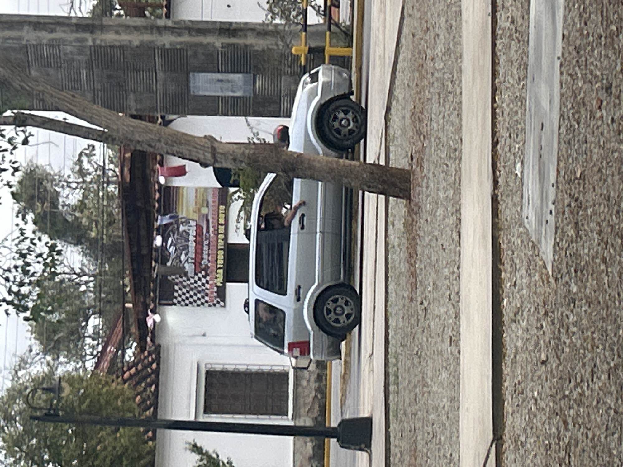A silver Ford EcoSport is parked in front of a white building with a tiled roof. The building has a large checkered banner hanging on it. The car has its rear passenger-side tire deflated. A person's arm is visible out the driver's side window. The scene appears to be in an urban or suburban area, possibly Santa Ana, Venezuela, given the building's architecture and the surrounding vegetation. A street lamp is visible to the left of the car, and a tree trunk is in the foreground to the right.