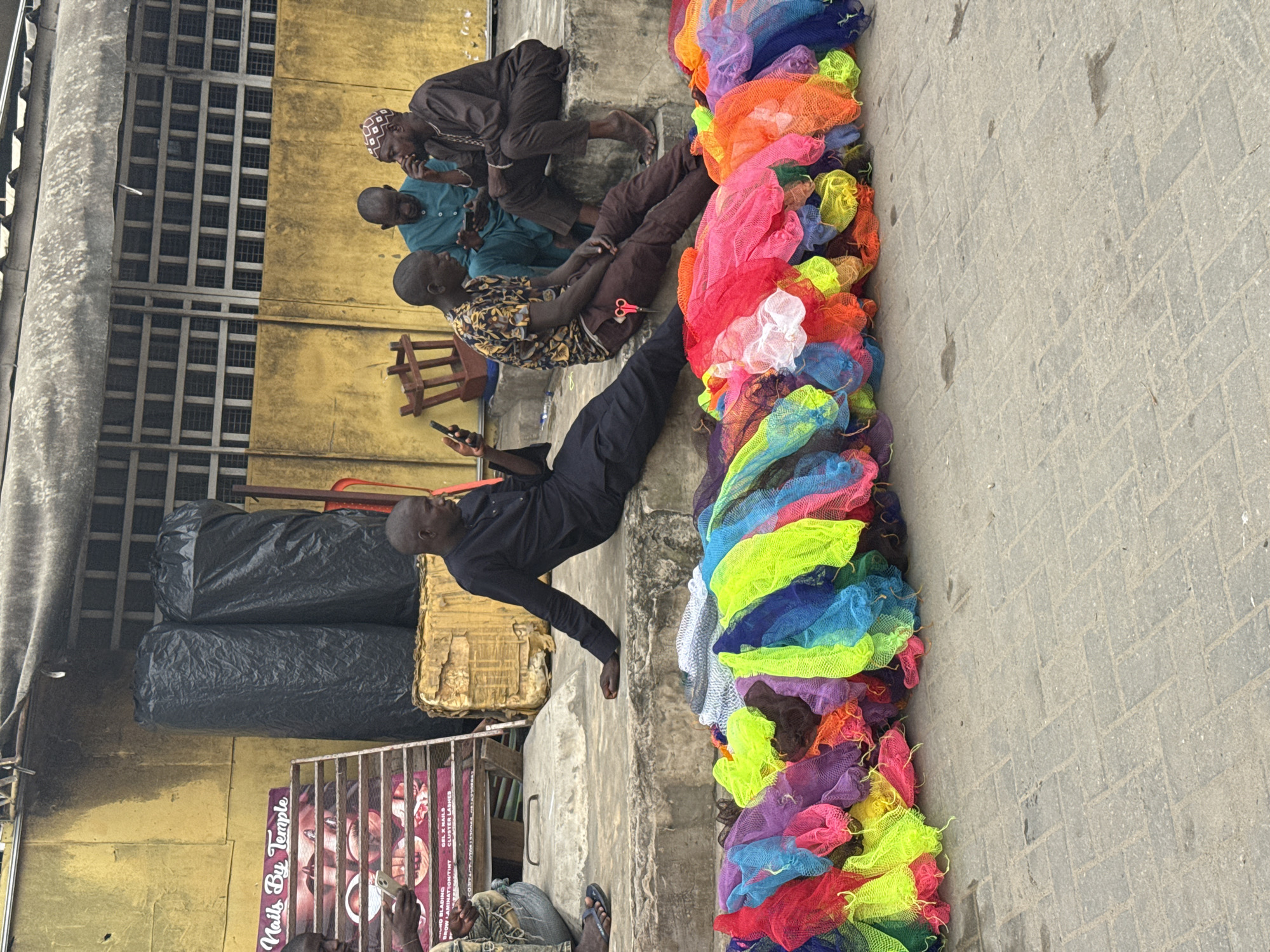 The image depicts a street scene in Satellite Town, Nigeria, where four men are casually sitting on a concrete ledge in front of a yellow-walled building. In the foreground, a vibrant collection of mesh bags in various colors – red, pink, yellow, green, blue, purple, and orange – is neatly arranged.
Two men are sitting on the ledge, facing each other. One is wearing a dark outfit and looking at his phone, while the other, in a patterned short-sleeved shirt and brown trousers, has a pair of scissors tucked into his pocket. Further to the right, two other men are seated. One, in a blue traditional garment, looks down at his phone. The man to his right, dressed in a dark brown traditional outfit and a patterned cap, appears to be in conversation, holding his hand to his face.
Behind the men, the building's facade is a striking yellow, with a row of barred windows above. To the left, a metal rack displays posters advertising "Nails By Temple," with images of nails and beauty treatments. Two large, black, cylindrical objects, possibly covered goods or trash bags, are visible next to the rack. The ground in front of the ledge is paved with interlocking concrete blocks.