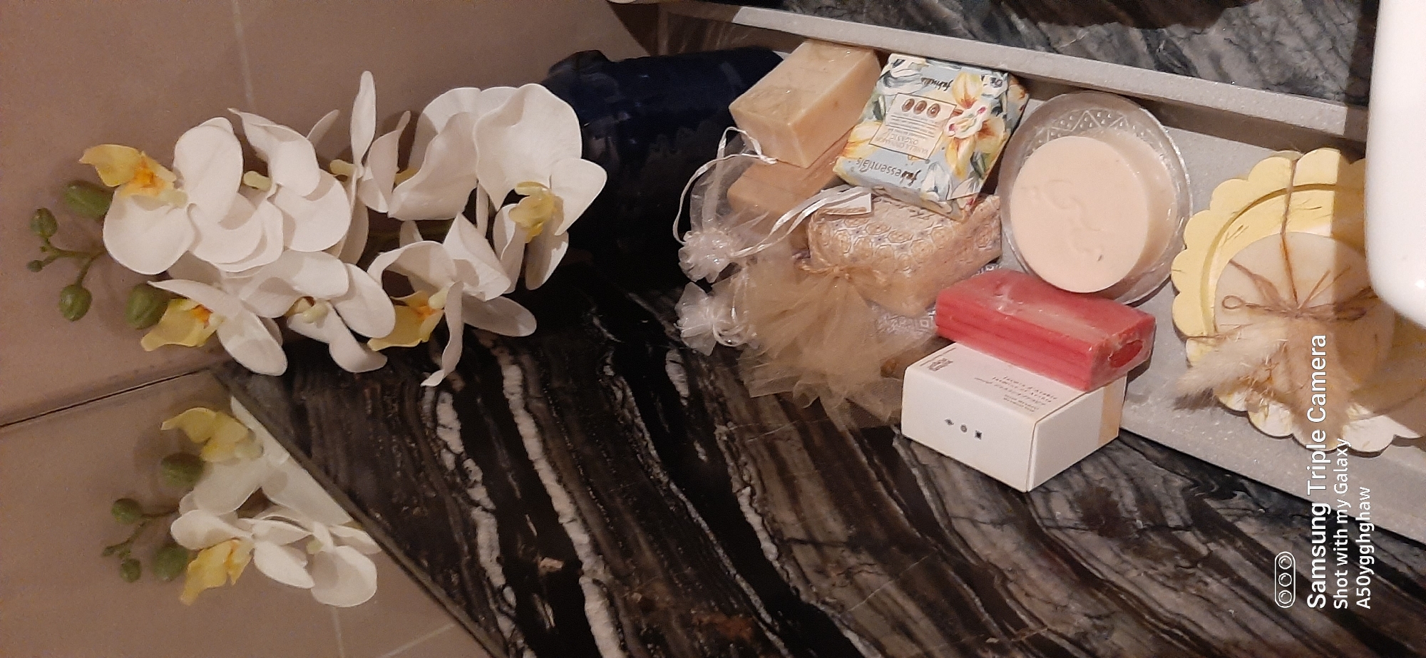 This is a close-up, eye-level shot of a collection of bath products and artificial orchids, likely arranged in a bathroom. The items are placed on a marble surface with a dark, swirled pattern and are partially contained within a light grey tray.
In the foreground, there is a white box with a red rectangular object next to it, possibly a bar of soap or bath bomb. To the right of these, a circular, light-colored soap rests on a clear, decorative glass plate. Further to the right, another circular item, also appearing to be soap, is decorated with a piece of dried grass and twine. In the background, several small, sheer bags with drawstring closures contain what appear to be bath salts or small soaps. Two larger, rectangular bars of soap are also visible, one wrapped in floral paper and another plain tan bar.
Above and behind the bath products, artificial white orchids with yellow accents are arranged, with some green buds visible. The overall impression is one of a curated display of bath and body items, possibly for a spa experience or gift.
The image is a photograph taken with a Samsung Triple Camera, shot with a Galaxy A50yggghaw. There is no indication of the specific location within the city of Dubai, United Arab Emirates, or any people or actions occurring. The scene is static and appears to be an indoor setting.