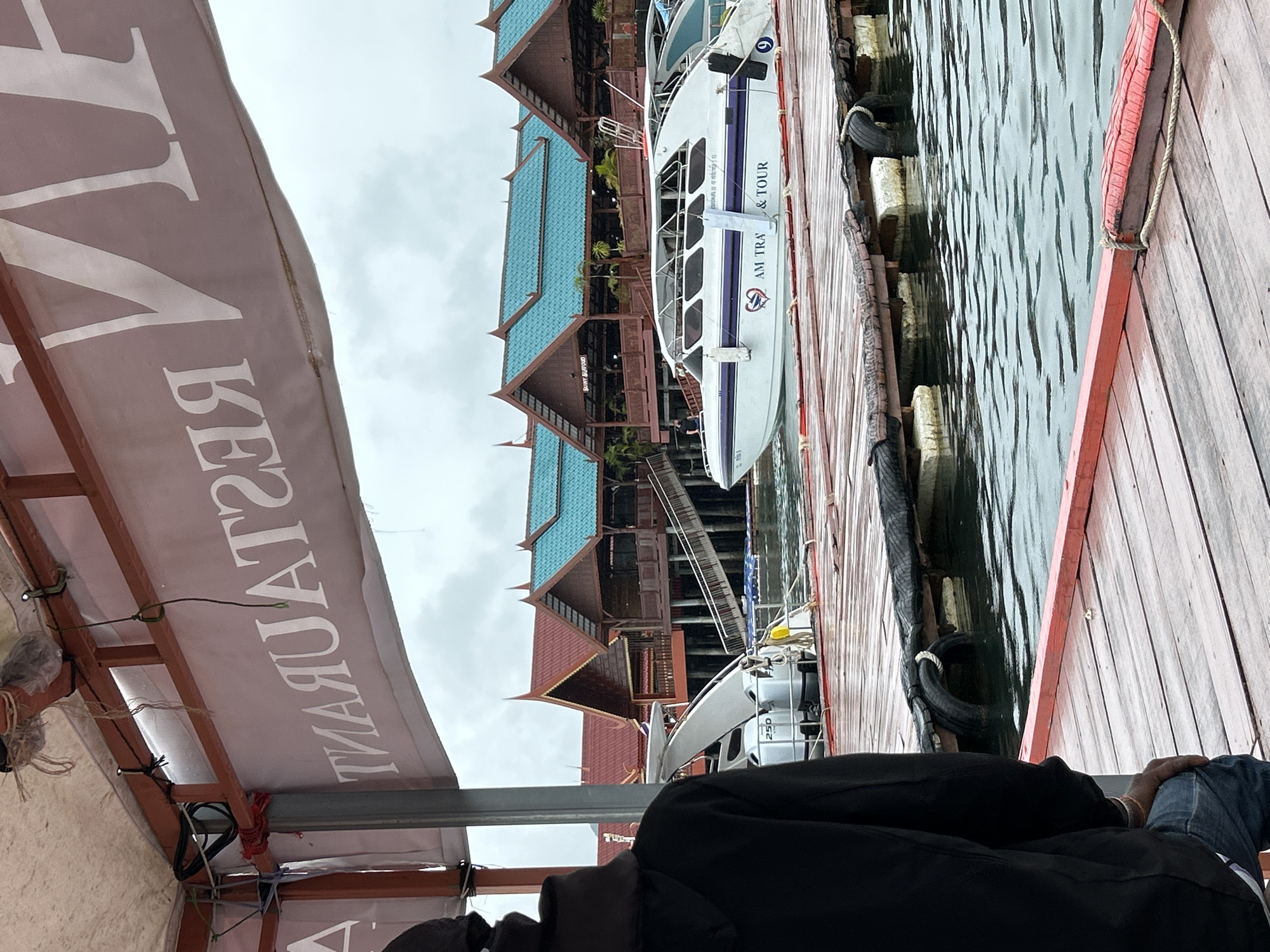 The image shows a view from a boat in Ko Panyi, Thailand. In the foreground, a person wearing a black hoodie and blue jeans is seen from behind, sitting on a wooden dock. To the left, the back of a boat with "250" written on it is partially visible. A white speedboat with "AM TRA & TOUR" written on its side is docked to the right. Behind the speedboat, several wooden buildings with distinctive turquoise roofs and red trim are situated on stilts over the water. A large banner with white text, which appears to read "RESTAURANT" in reverse, is visible in the upper portion of the image, attached to the structure of the boat from which the photo is taken. The water is calm, and the sky is overcast.