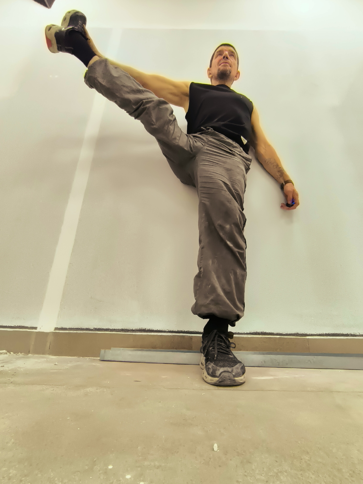 A man in athletic attire is performing a leg split against a white wall. He is positioned with his left leg extended vertically upwards, supported by the wall, while his right leg is bent at the knee and planted on the floor. He wears a black sleeveless shirt, grey pants, black socks, and athletic shoes. The environment appears to be an indoor space with a textured floor and some construction materials, including metal strips along the base of the wall. The lighting is bright, casting subtle shadows. The man's expression suggests focus and concentration. The image does not provide any specific information about the location being the city of Qarağandı, Kazakhstan.