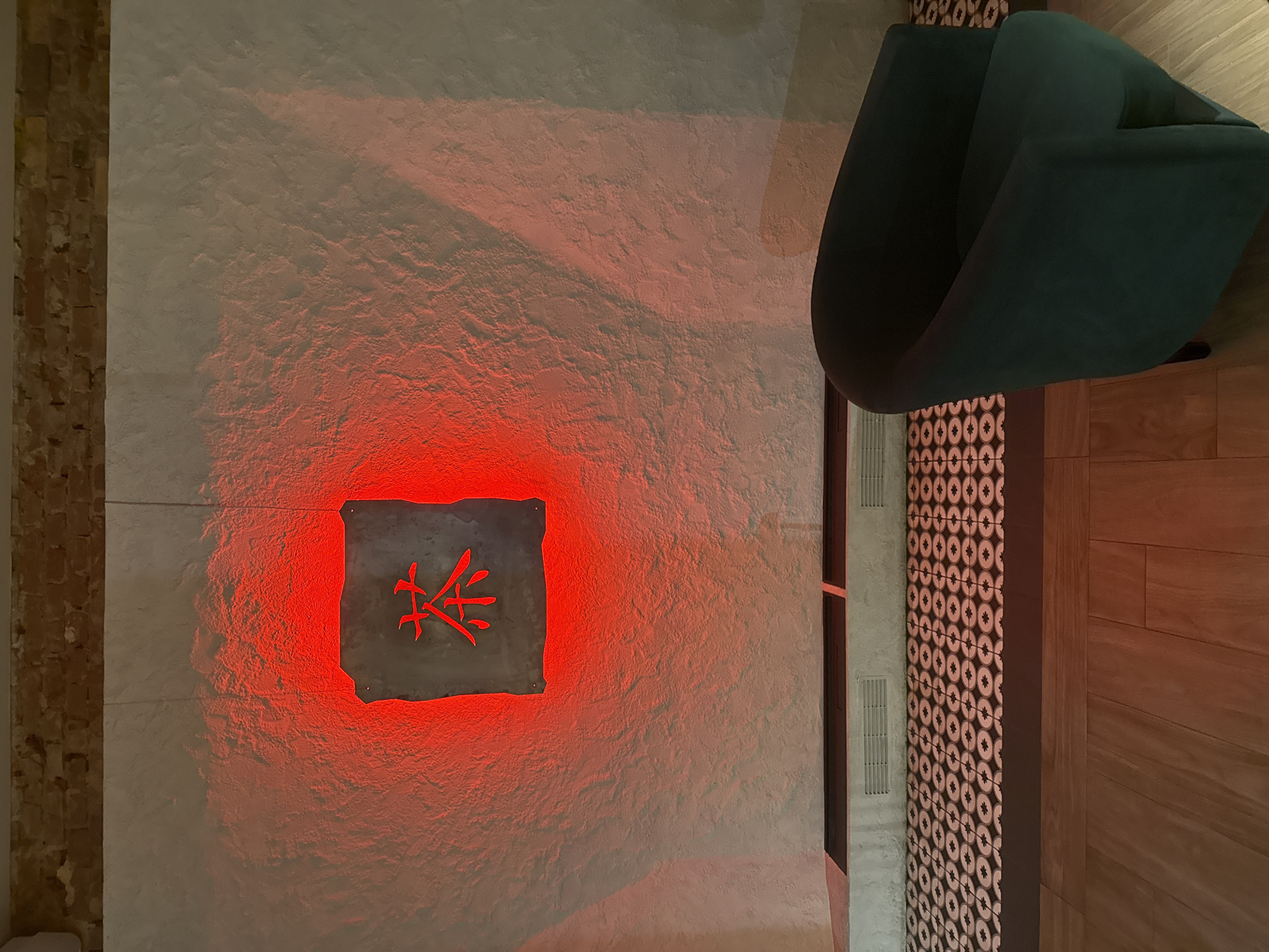 An interior scene in Unknown, Unknown, features a textured light-colored wall with significant red illumination emanating from behind a central wall-mounted plaque. The plaque is dark, rectangular with an irregular edge, and contains a red-lit Chinese character "茶" (chá, meaning tea) cut into its surface. Above the textured wall, a band of exposed red brickwork is visible. Below the textured wall, a low, light-colored structure with multiple rectangular vents spans horizontally. The floor transitions from a section of dark tiles with a repeating light circular pattern to light-brown wooden planks in the foreground. A dark green, curved armchair is positioned on the right side of the wooden floor, angled towards the viewer. No individuals are present.