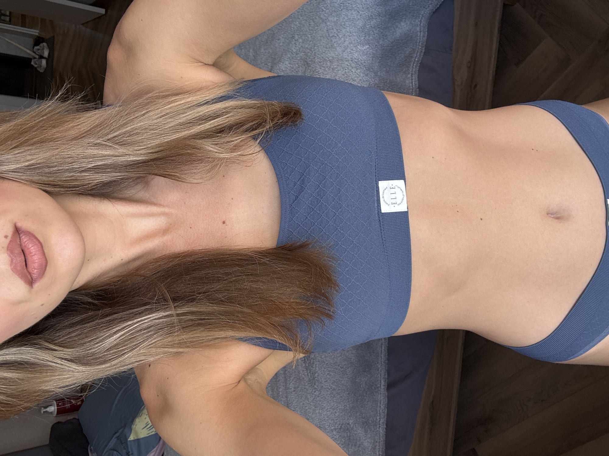 The image shows a woman wearing a matching blue sports bra and underwear set. She is lying down on a gray surface, possibly a bed or couch. The photograph is taken from a high angle, looking down on her. Her hair is blonde with darker roots. The brand label on the sports bra is visible. The background includes a glimpse of a wooden floor and a bedside table. The location is Unknown, Unknown.
