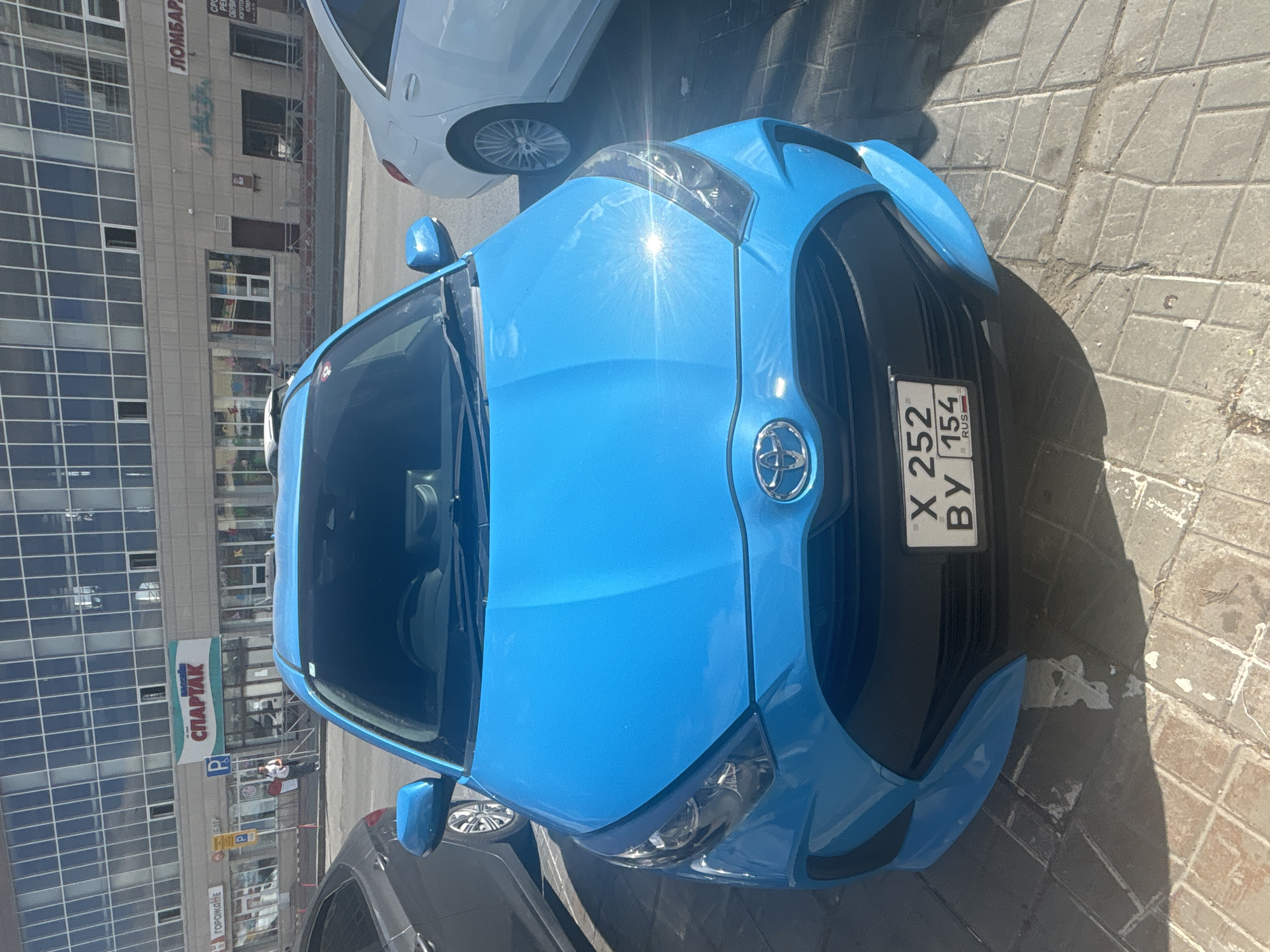 A bright blue Toyota Yaris is parked on a paved area in front of a building with large glass windows, likely a commercial establishment, in Novosibirsk, Russia. The building has signage that includes Russian text such as "СПАРТАК" and "ЛОМБАРД". A white sedan is parked to the right of the Yaris, and a dark grey vehicle is partially visible to the left. A person is walking on the sidewalk in front of the building. The license plate of the Toyota Yaris reads "X 252 BY 154 RUS". The scene is illuminated by bright sunlight, casting distinct shadows on the ground.