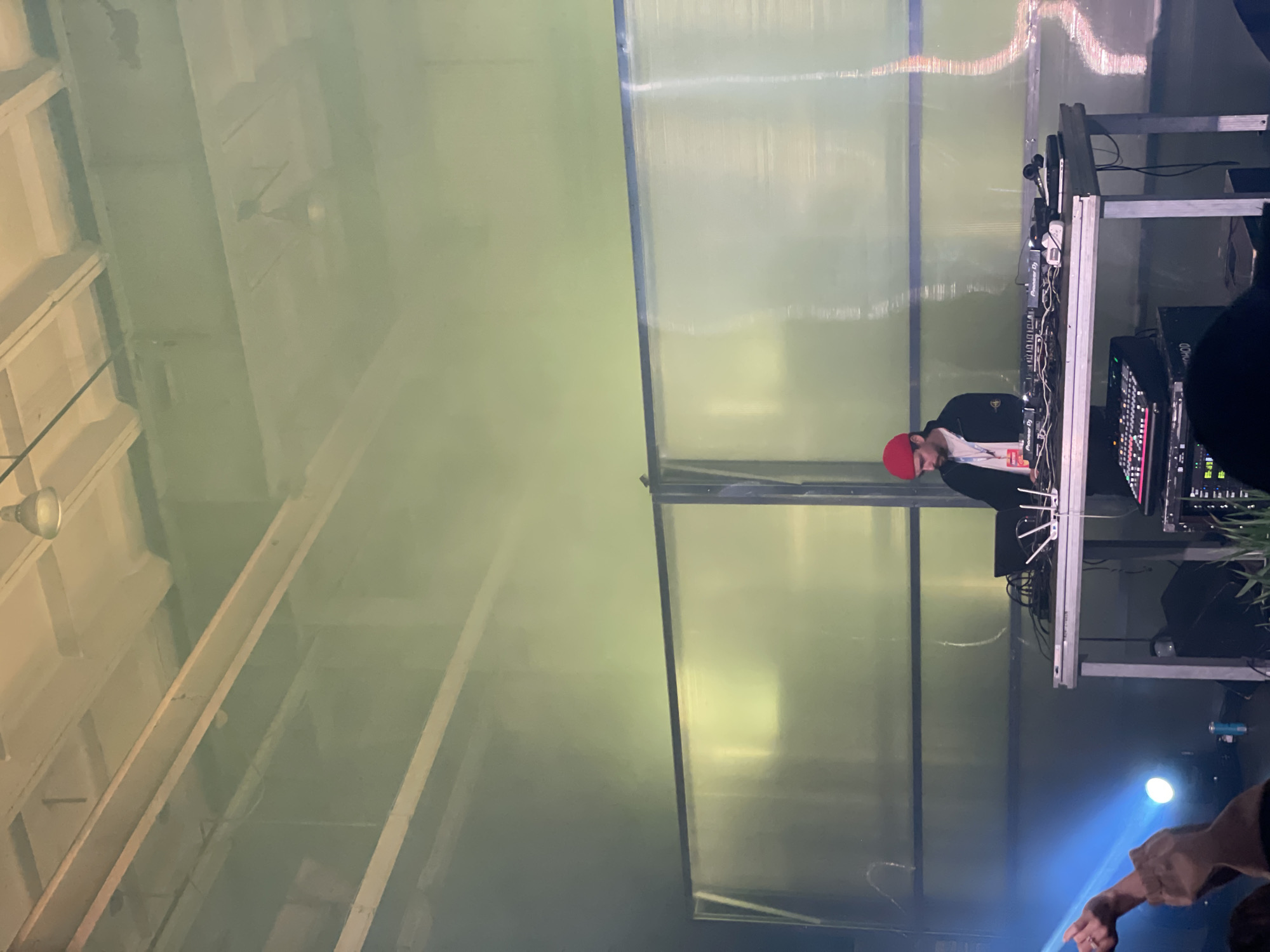 A DJ with a red cap is performing behind a mixing console at a venue. The console is on a metal stand, and behind the DJ is a translucent wall with metal framing. The environment appears to be indoors, possibly a warehouse or large event space with metal framing in the ceiling. The location is likely in Saint Petersburg, Russia.
