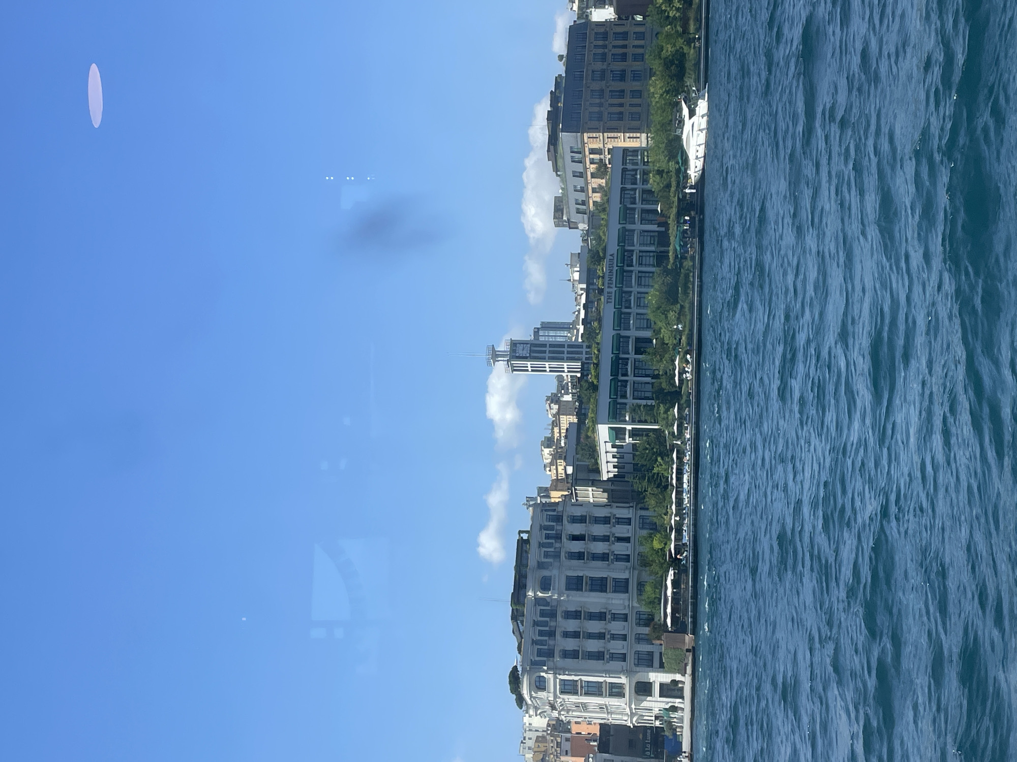 A wide shot captures the Bosphorus Strait in Istanbul, Turkey, with a view of the Beyoğlu district skyline under a clear blue sky with scattered white clouds. The water in the foreground is a deep blue, with visible ripples indicating movement. Along the shoreline, several buildings are visible, including a prominent white multi-story building with many windows and a tiered roof, and a longer, lower green-roofed building with the text "THE PENINSULA" clearly displayed. Behind these, other buildings of varying architectural styles and colors rise up the hillside. A white yacht is moored near the shore. The overall scene depicts a sunny day in a bustling urban waterfront area.