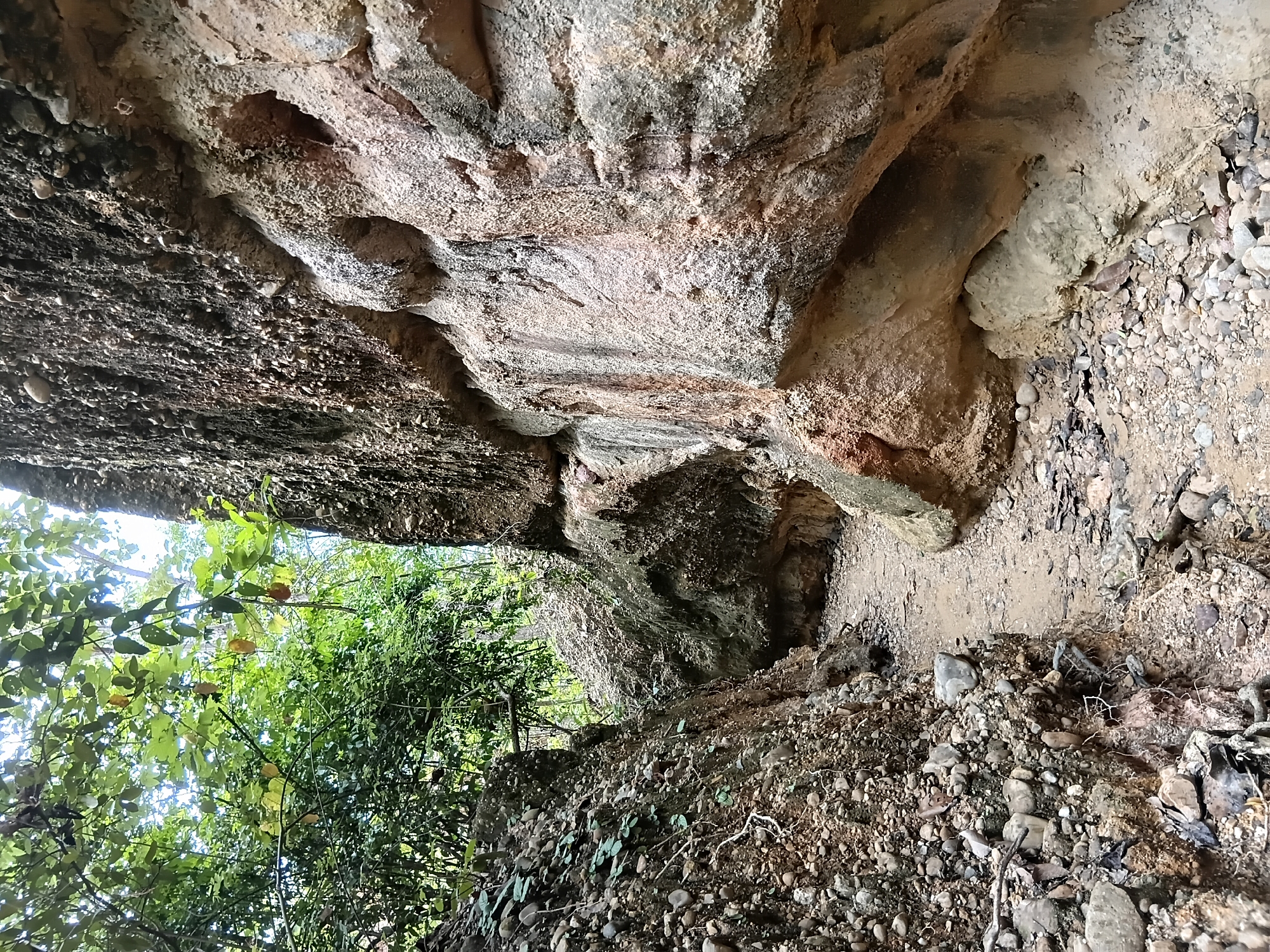 The image depicts a narrow, winding path through a gorge with steep, rocky walls. The path is covered in dirt and small pebbles. The left side of the gorge is characterized by a wall of loose rocks and soil, with some green foliage growing from it. The right side features a more solid rock formation with visible layers and some erosion, creating indentations and overhangs. The upper parts of the gorge walls are obscured by dense green foliage and tree branches, with some of the leaves appearing yellowish, suggesting they might be fruit-bearing or nearing a change in season. The sky is visible through the canopy in some areas, appearing bright and possibly overcast. The overall impression is of a natural, somewhat rugged environment, likely a hiking trail or natural feature within a forested area. The location is identified as Ban Sop Hat, Thailand. There are no people or animals visible in the image.