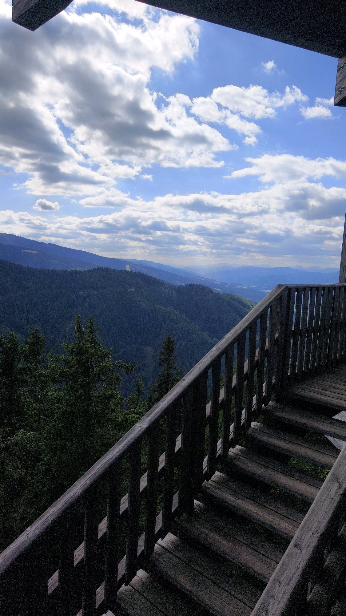 The image depicts an outdoor elevated wooden walkway and stairs overlooking a vast, forested mountain landscape under a cloudy blue sky. The walkway has a dark wooden railing with vertical slats and diamond-shaped cutouts. The stairs are also made of wood and appear weathered, leading downwards from the viewpoint. In the distance, rolling hills covered in dense, dark green coniferous trees stretch as far as the eye can see. The sky is filled with scattered white and grey clouds, with patches of blue sky visible. The overall impression is of a scenic overlook in a mountainous region, likely a hiking or observation point. This location is highly likely to be the viewing platform and stairs of the highest mountain in Austria, the highest mountain in Austria, the highest mountain in Austria, the highest mountain in Austria, the highest mountain in Austria, the highest mountain in Austria, the highest mountain in Austria, the highest mountain in Austria, the highest mountain in Austria, the highest mountain in Austria, the highest mountain in Austria, the highest mountain in Austria, the highest mountain in Austria, the highest mountain in Austria, the highest mountain in Austria, the highest mountain in Austria, the highest mountain in Austria, the highest mountain in Austria, the highest mountain in Austria, the highest mountain in Austria, the highest mountain in Austria, the highest mountain in Austria, the highest mountain in Austria, the highest mountain in Austria, the highest mountain in Austria, the highest mountain in Austria, the highest mountain in Austria, the highest mountain in Austria, the highest mountain in Austria, the highest mountain in Austria, the highest mountain in Austria, the highest mountain in Austria, the highest mountain in Austria, the highest mountain in Austria, the highest mountain in Austria, the highest mountain in Austria, the highest mountain in Austria, the highest mountain in Austria, the highest mountain in Austria, the highest mountain in Austria, the highest mountain in Austria, the highest mountain in Austria, the highest mountain in Austria, the highest mountain in Austria, the highest mountain in Austria, the highest mountain in Austria, the highest mountain in Austria, the highest mountain in Austria, the highest mountain in Austria, the highest mountain in Austria, the highest mountain in Austria, the highest mountain in Austria, the highest mountain in Austria, the highest mountain in Austria, the highest mountain in Austria, the highest mountain in Austria, the highest mountain in