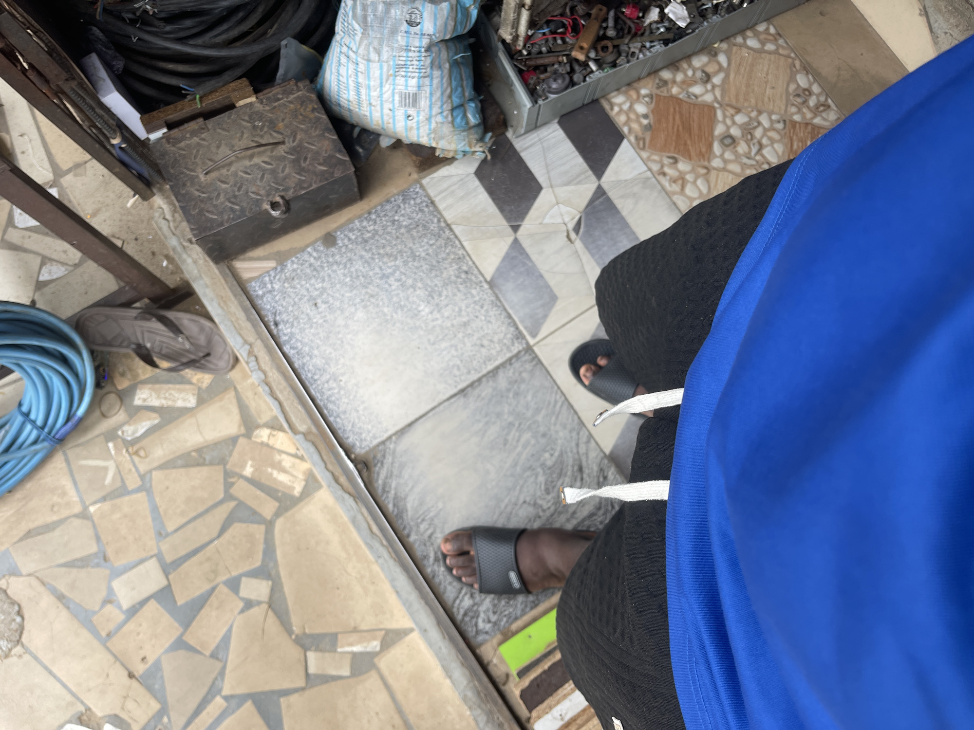 A high-angle, overhead shot shows a person's legs and feet from the knees down, standing on a tiled outdoor surface. The person is wearing black shorts with white drawstrings, and a bright blue t-shirt. They are wearing black slide sandals on both feet. The ground consists of various tiles, including broken, irregular beige ones, square gray and white marbled tiles, and patterned beige and brown tiles. To the right of the person's feet, there is a metal toolbox, a plastic bag with blue and white stripes, and a tray filled with various metal parts and tools. In the background, a blue hose is coiled up, and a brown flip-flop is visible on the tiled ground. A metal grate and some electrical wires are also present.