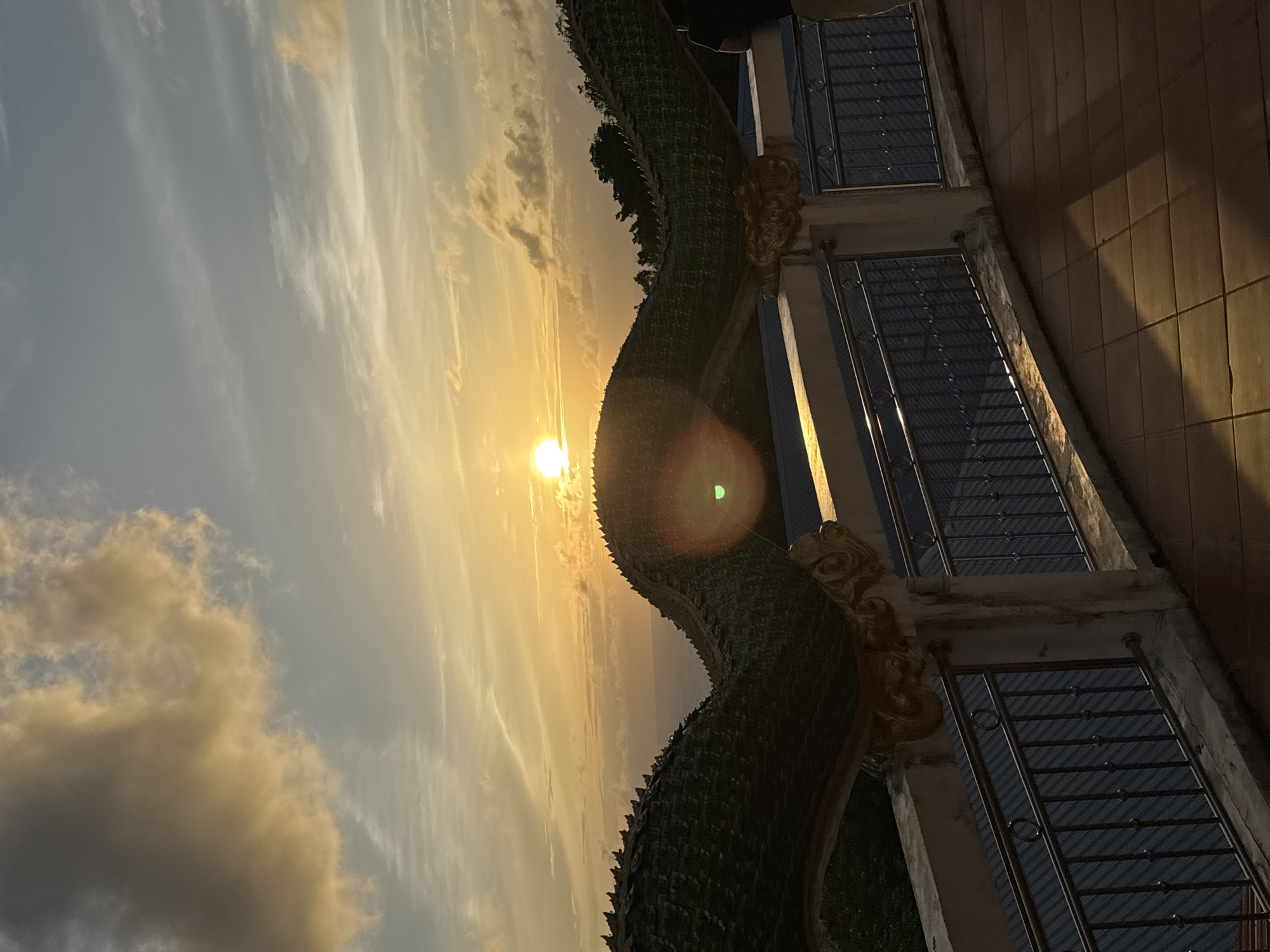 The image captures a sunset over the ocean, viewed from a balcony with a railing. In the foreground, there is a large, ornate structure resembling a dragon's body, with a textured, dark green surface and golden decorative elements. The balcony floor is paved with square tiles. In the background, the sky is filled with dramatic clouds illuminated by the setting sun, casting a warm golden glow. The ocean is visible on the horizon. This scene is likely located at a viewpoint in Patong, Thailand, possibly at a temple or a scenic overlook offering panoramic views of the coast. There are no people prominently featured in the image, though a person's dark clothing is visible on the far right edge. The overall atmosphere is serene and picturesque, highlighting the natural beauty of the sunset.