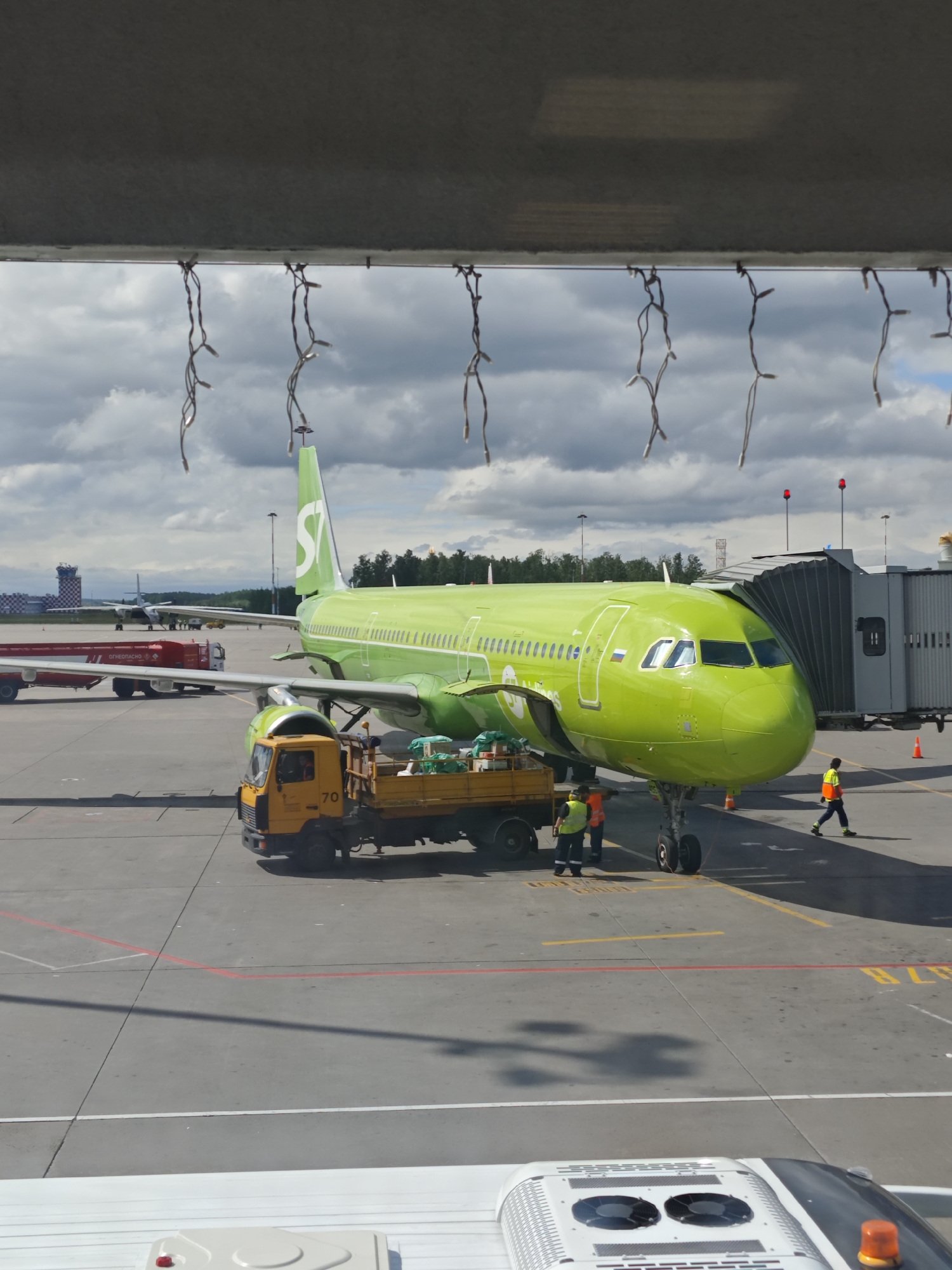 An S7 Airlines airplane, painted bright green with a white logo, is parked at Pulkovo Airport in Saint Petersburg, Russia. The aircraft is connected to a jet bridge. A yellow service truck with the number "70" on its side is parked next to the airplane's landing gear. Two airport workers in high-visibility vests are standing near the truck. Another worker in an orange vest is walking away from the airplane towards the right side of the image. In the background, another airplane is visible, along with airport control towers and trees. The sky is partly cloudy. The scene appears to be a typical day at the airport, with ground operations underway.