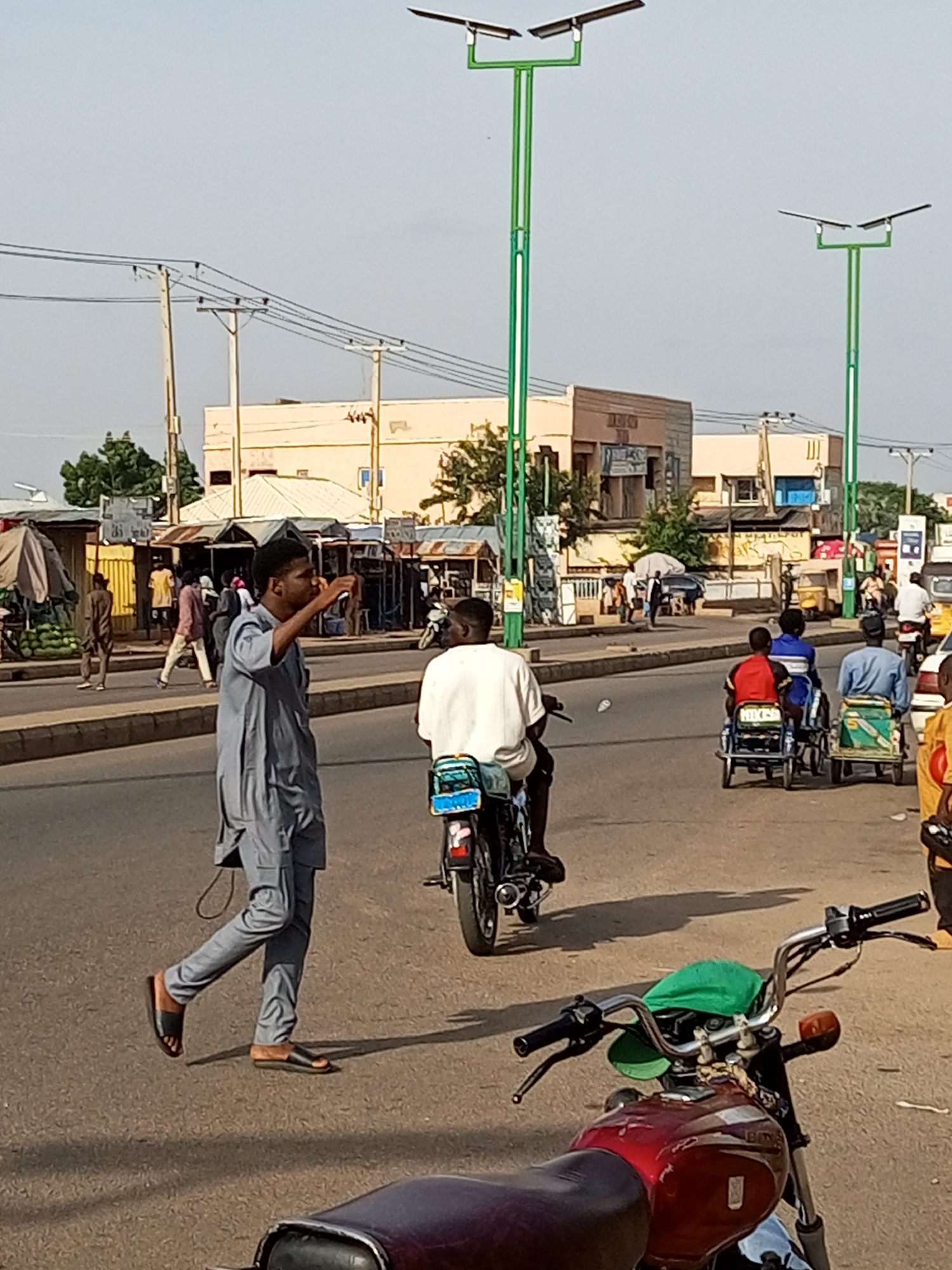 This image depicts a street scene with several people and vehicles under a clear sky, suggesting daytime. The setting appears to be a market or busy commercial area with buildings lining the street, some appearing to be shops.
In the foreground, a man in a grey traditional outfit is walking and holding up a phone, possibly taking a picture or making a call. Another man is riding a motorcycle behind him, also wearing a white shirt. Further down the street, several people are seen in a motorcycle-drawn cart, with one person in front driving and two passengers behind. Other individuals are visible in the background, some walking and others on motorcycles.
Two prominent streetlights with solar panels are visible in the midground, indicating a modern infrastructure in what could be an urban or semi-urban area. The weather seems to be warm and sunny, with distinct shadows cast by the people and objects.
The overall atmosphere suggests a bustling environment, possibly during the day's activities. There is no visible text in the image that provides specific location information or other context.