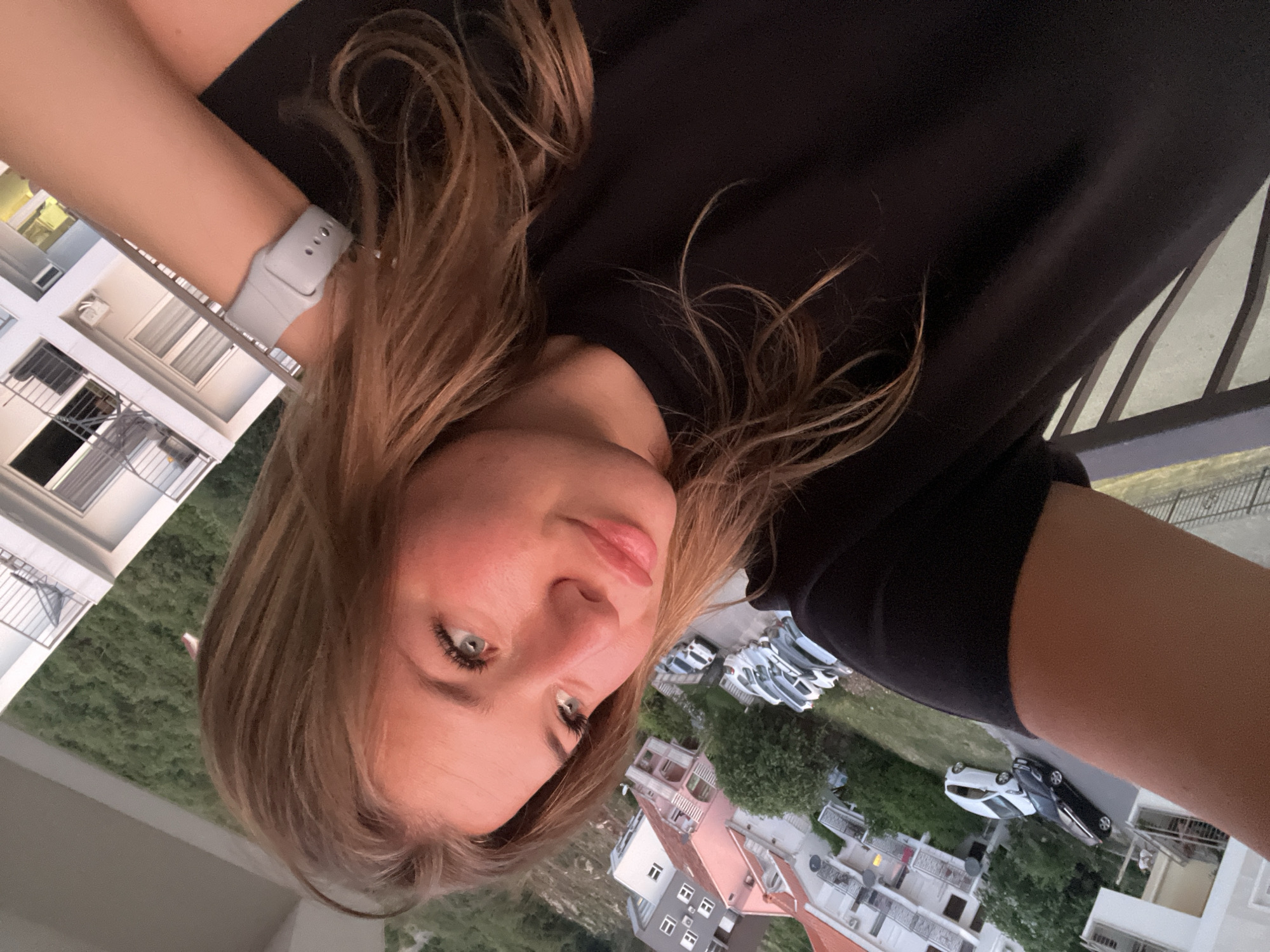 A young woman with long brown hair and blue eyes is taking a selfie. She is wearing a black t-shirt and a grey smartwatch. The background shows a street with parked cars and buildings with white walls and red roofs. The scene appears to be in a residential area of Budva, Montenegro, as suggested by the architecture and landscape.