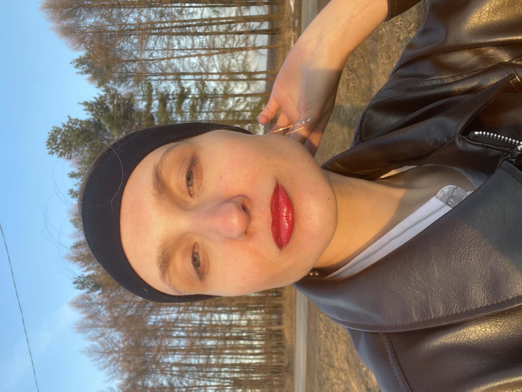 The image shows a selfie of a woman wearing a black headband and a black leather jacket over a white t-shirt. Her right hand is raised near her head, and she is looking directly at the camera. She has fair skin, light eyes, and red lipstick. The background features a line of trees and a clear, light blue sky. Based on the presence of Подшивалово in the query, it can be assumed that the photograph was taken in the city of Podshivalovo, Russia. The scene appears to be outdoors.