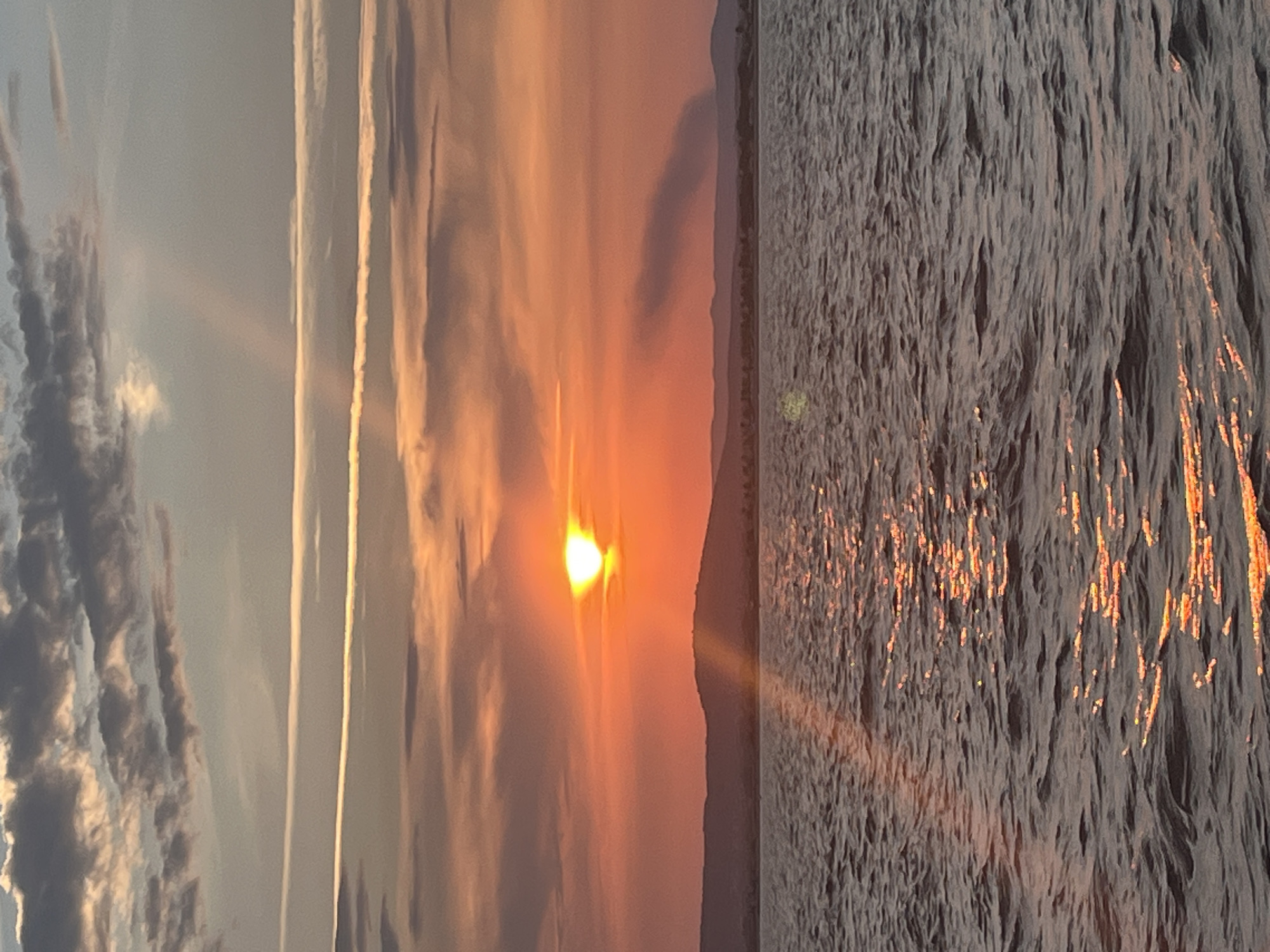This image depicts a sunset over the Mediterranean Sea, viewed from Palavas-les-Flots, France. The sun, a bright orange orb, is setting near the horizon, casting a warm glow across the sky and reflecting on the rippling water. The sky is filled with a mix of clouds, some tinged with orange and pink from the sunset, while others are darker grey. Two distinct contrails are visible in the upper sky, suggesting air traffic. A faint, dark plume of smoke rises from the distant shoreline, which is marked by a silhouetted, hilly landscape. The water appears choppy, with waves catching the sunlight, creating a shimmering effect. No people or specific landmarks of Palavas-les-Flots are clearly identifiable in this broad seascape.