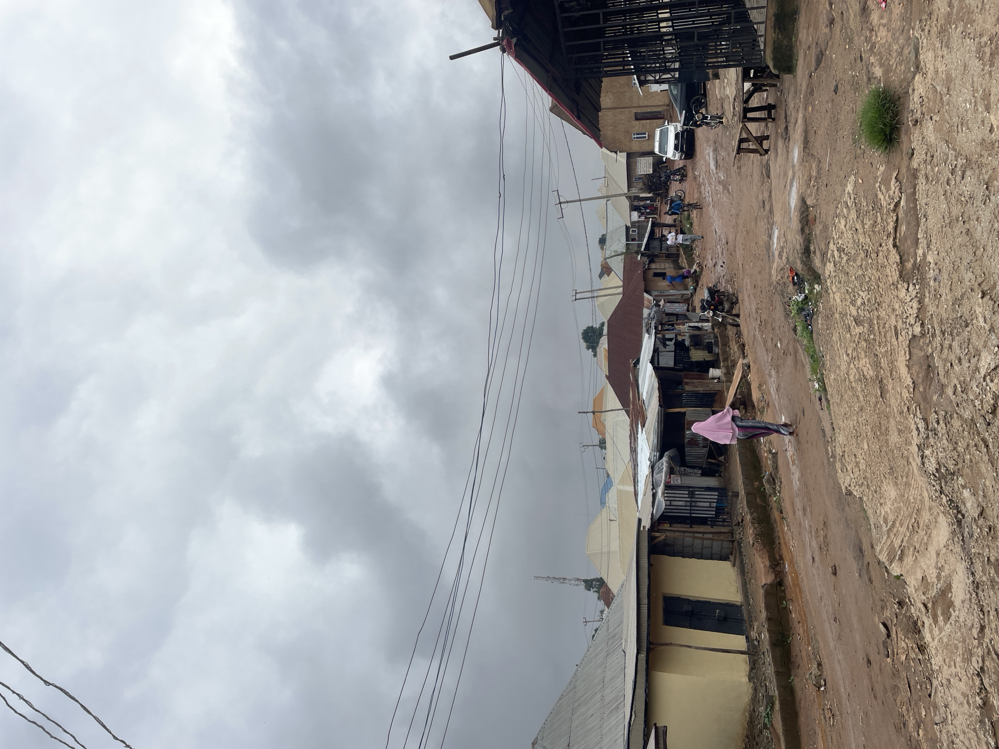 The image depicts a street scene in Kurunduma, Nigeria, under a heavily overcast sky. The unpaved road is muddy and uneven, showing signs of recent rain with puddles and erosion. On either side of the road are buildings with corrugated metal roofs.
In the foreground, a person wearing a pink headscarf and a patterned skirt walks away from the camera down the street. To the left, a yellow building with a dark door is visible. Further down the street, several other structures are present, some appearing to be shops or residences with open fronts.
Several motorcycles are parked along the road. A white SUV is parked on the right side of the street, and a black car is partially visible behind it. A wooden bench or table is situated on the right side of the road in front of a building with a metal fence.
In the distance, a cell phone tower can be seen rising above the rooftops. The overall impression is a typical day in a residential and commercial area of Kurunduma, experiencing inclement weather.