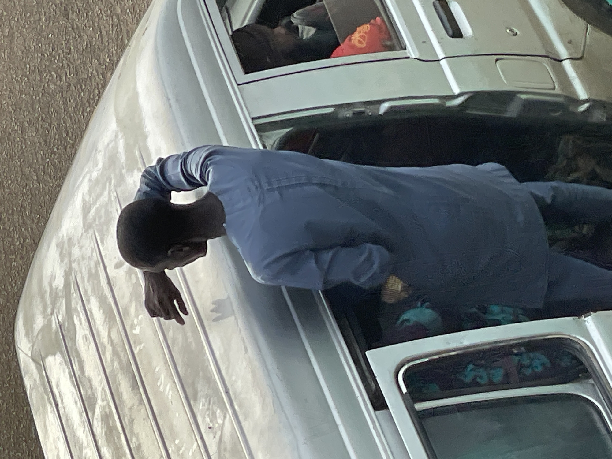 An overhead shot captures a man standing next to the open door of a silver van. He is dressed in a blue tunic and pants and is looking away from the camera, with his right arm raised and hand touching his forehead. Inside the van, two figures are visible. One person, wearing a dark head covering, is seated in the back, and a red object, possibly a bag or cloth, is near them. Another person, partially obscured, is seated in the middle row. The van is parked on a textured, light brown surface, which appears to be a road or ground.