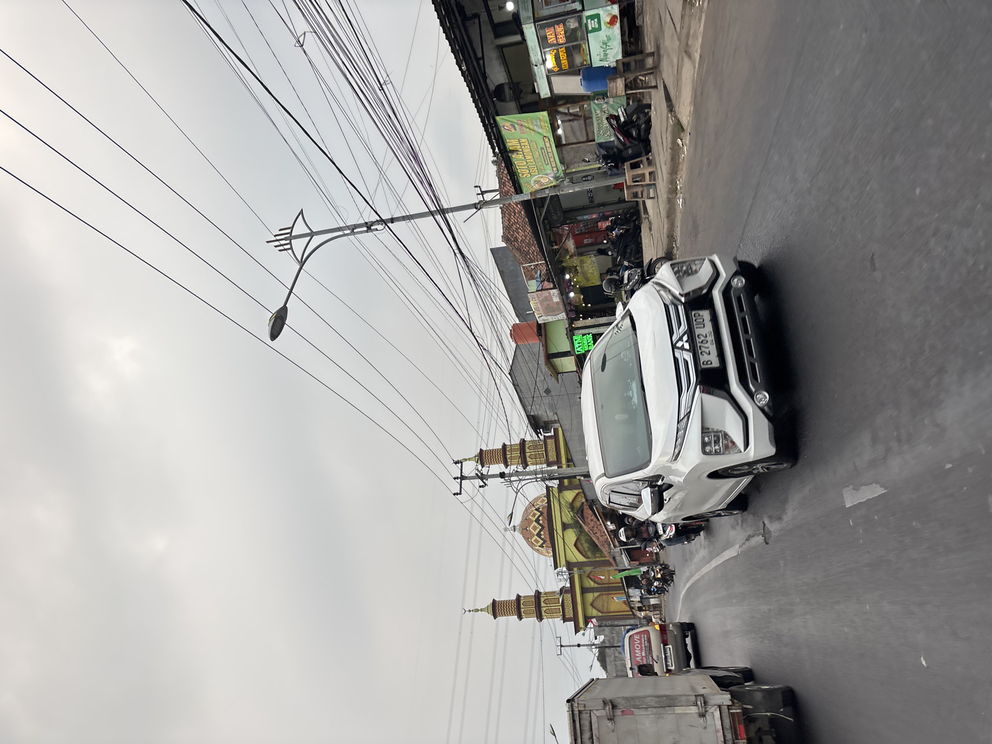 The image depicts a street scene in Bekasi, Indonesia, on an overcast day. A white SUV, identified by its license plate B 2762 UOP, is prominently in the foreground, driving on a paved road. To the left of the SUV, a white truck is also visible. Several motorcycles are on the road, some carrying riders. In the background, a mosque with a dome and two minarets stands out. Adjacent to the mosque are several small buildings, some of which appear to be food stalls or shops, with signage visible, including one with text in Indonesian that reads "SOTO & AYAM ASLI LAMONGAN." Overhead, a complex network of electrical wires is strung across the scene, supported by utility poles, one of which has a street lamp. The general environment appears to be a busy urban or suburban area with traffic and commercial activity.
