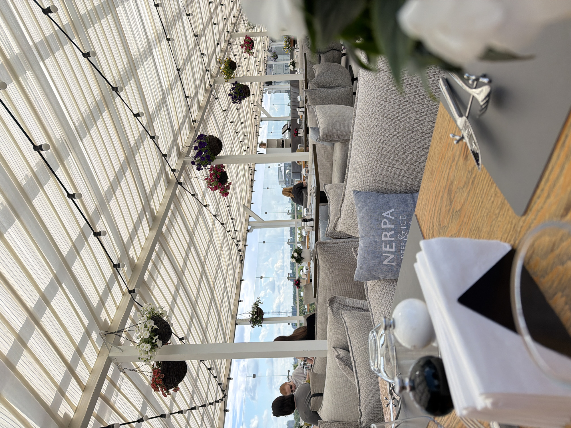 The image depicts a rooftop restaurant in Saint Petersburg, Russia, with an open-air dining area covered by a translucent roof with string lights. The setting appears to be a daytime scene, with a cloudy sky visible beyond a glass railing. Several tables are arranged with grey upholstered seating.
In the foreground, a wooden table is set with a white napkin holder, a salt and pepper shaker, and a glass. A fork and knife are also placed on a grey placemat. A light blue cushion with the word "NERPA" is visible on one of the seats.
In the middle ground, several patrons are seated at various tables. One group of three women is visible from behind, facing the view. Another woman is seated at a table further back.
Hanging baskets with flowers are suspended from the roof structure, adding a decorative element to the space. The overall ambiance suggests a modern and comfortable dining experience.