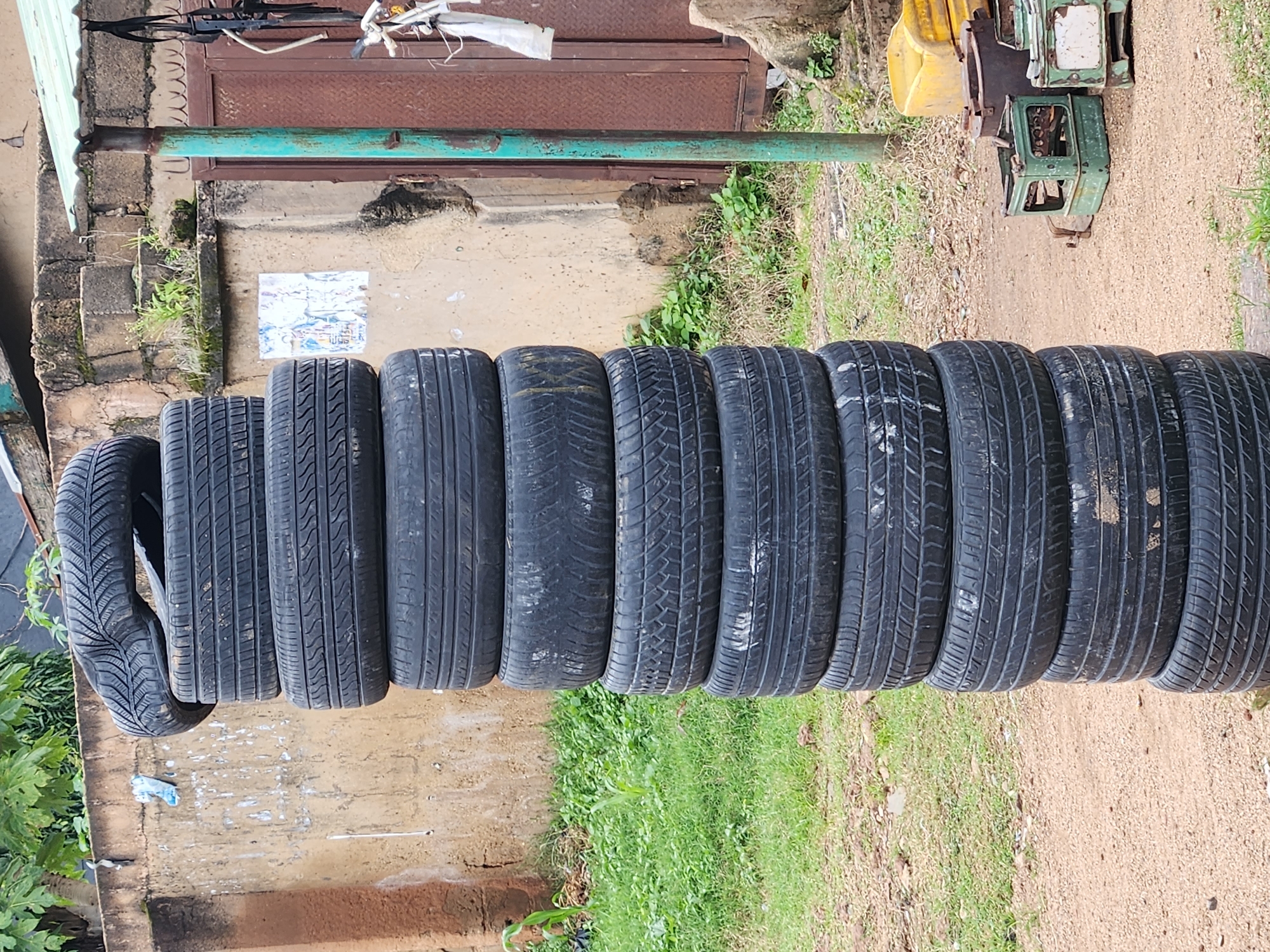 A stack of at least ten used car tires stands vertically in the foreground on Rukuba Road, Nigeria. The tires are black and show signs of wear. The background features a textured, light-brown concrete wall, overgrown green vegetation, and a rusty metal gate. To the right of the tires, there are various items including a yellow plastic container, a green metal crate, and a small white object. The ground is a mix of gravel and dirt. The image is captured from a slightly low angle, looking upwards towards the tires and the wall.