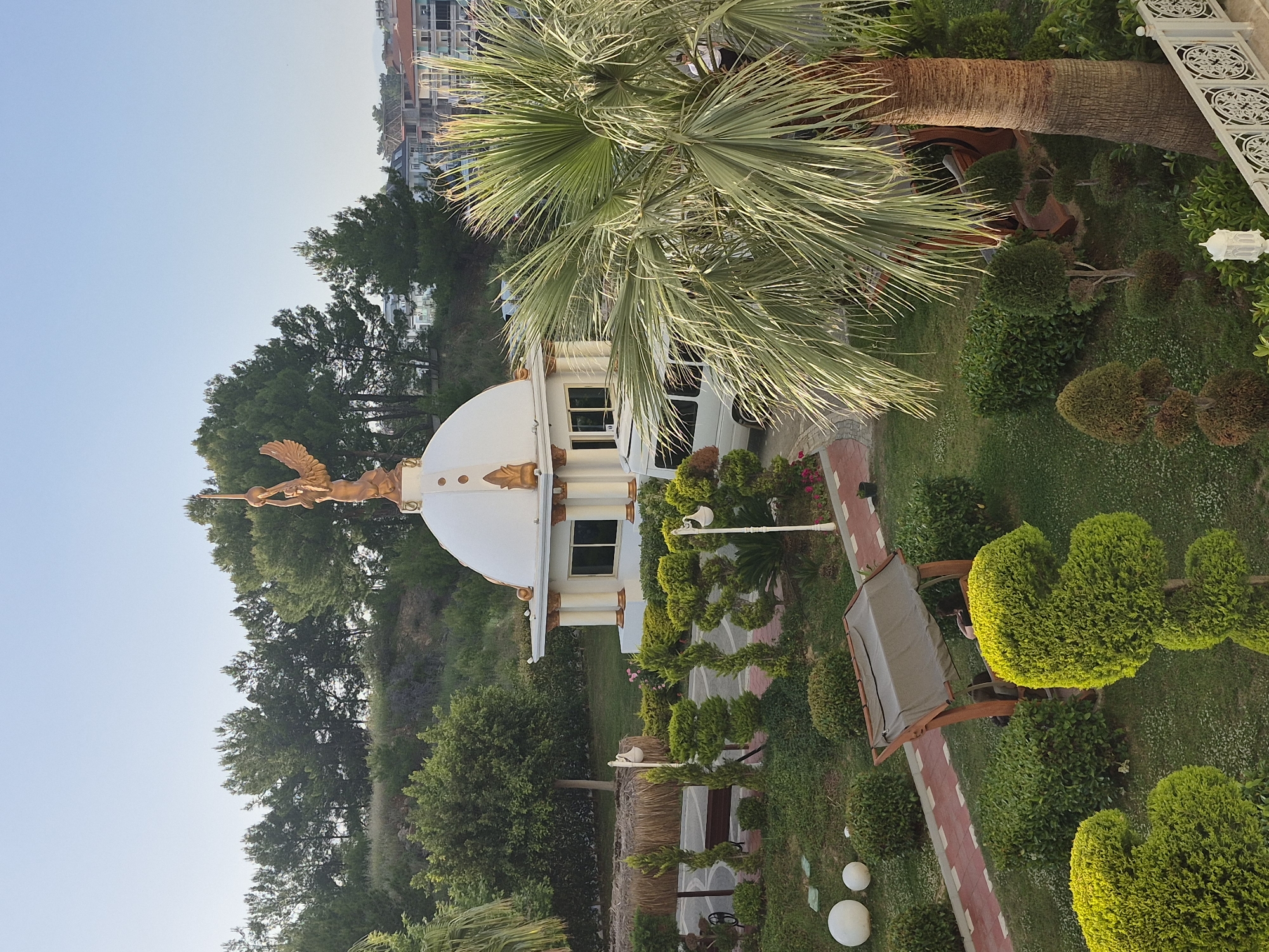 Here's a detailed description of the image:
**Scene:** A meticulously landscaped garden in Alanya, Turkey, featuring a prominent white domed structure with a golden statue atop it. The structure has windowed sides and decorative columns.
**Environment:** The garden is lush and well-maintained, with various types of trees, bushes, and manicured hedges, some trimmed into rounded shapes. A palm tree is visible, partially obscuring the structure. A red-paved pathway winds through the garden. There's a shaded seating area with a bench. Further back, there's a simple shelter structure. Buildings are visible in the distance beyond the trees, suggestive of the city setting.
**Objects:** The main structure is the domed building with the golden statue. Other objects include the garden furniture (bench), paved pathway, various plants and trees, lighting fixture, and the domed structure on top of the white building.
**Action:** The statue, likely representing a figure in mythology or a local emblem, is posed in a dynamic stance. The garden layout is designed for strolling and relaxation.
**Interactions:** The interaction is limited to the artistic impression of the statue and the design of the garden. No people are explicitly present, suggesting a serene or private setting.
**Location:** Alanya, Turkey, is inferred from the architecture and vegetation characteristic of the region.