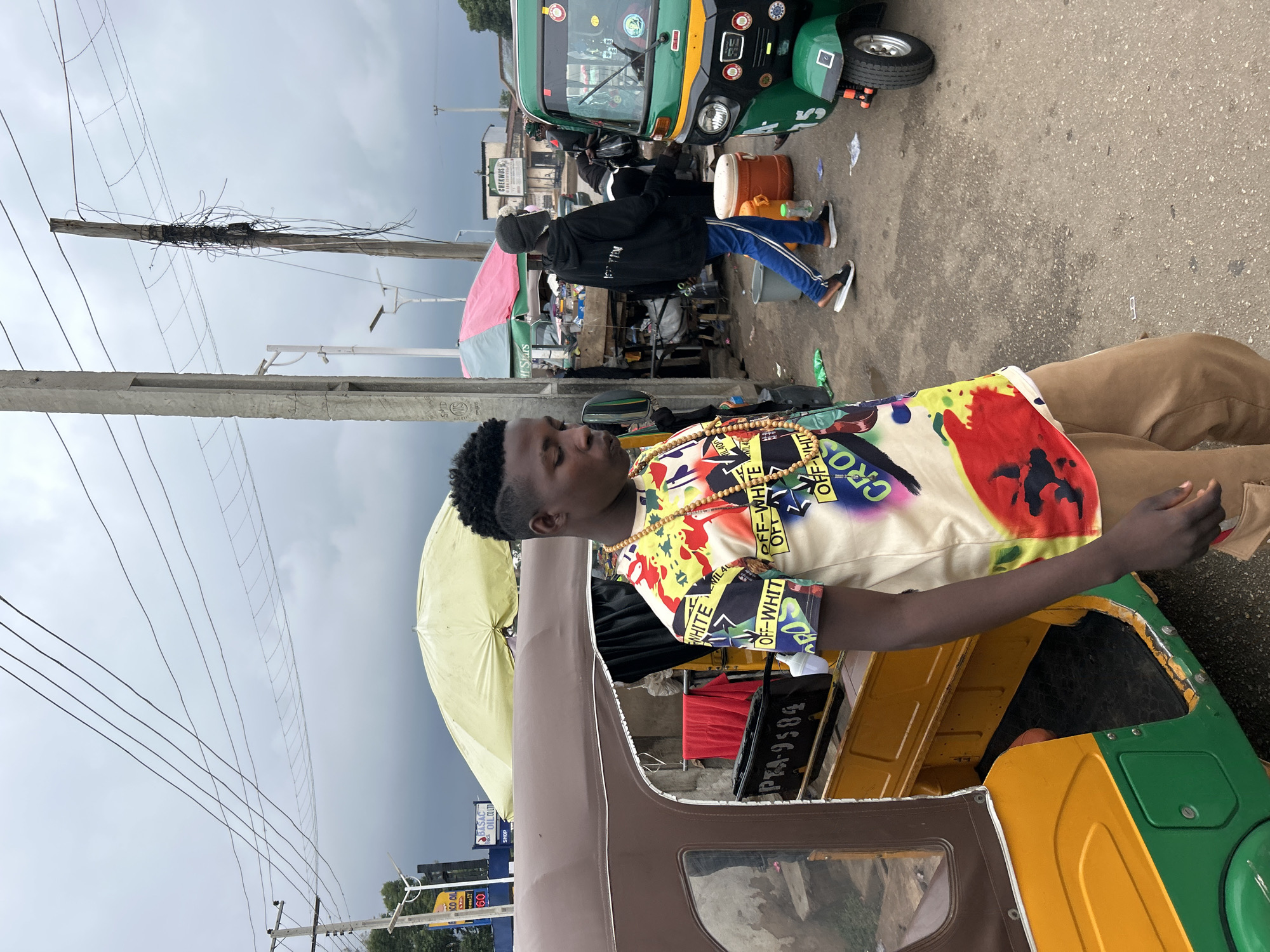 A young man stands in the foreground of a street scene in Jos, Nigeria, with a vibrant, colorful t-shirt. He is looking down and to his right. Behind him, a brown and yellow auto-rickshaw is parked, with the license plate "PTA-9584" visible. To the right of the auto-rickshaw, a green auto-rickshaw is also present. In the background, several people are walking, and market stalls with umbrellas are set up. The sky is overcast with dark clouds, suggesting an impending rain shower. The overall environment appears to be a busy street or market area.