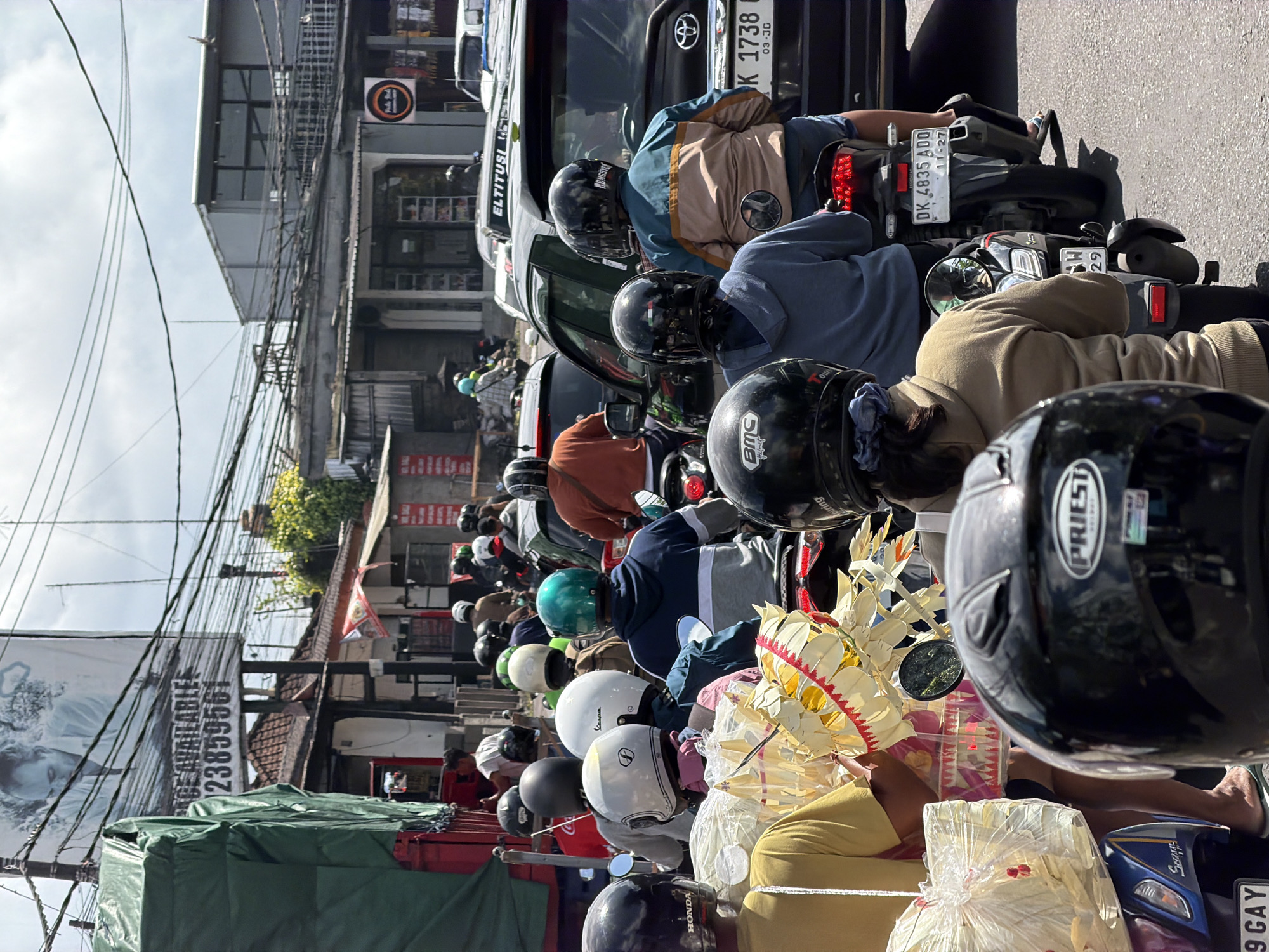 This is a medium shot of a traffic jam on a street in Mengwi, Indonesia. The scene is dominated by a line of motorbikes and some cars moving slowly along a paved road. Overhead, power lines crisscross the sky, and a large billboard with an image of a man and text advertising "SPACE AVAILABLE" is visible. Buildings line the street, some appearing to be shops with awnings.
The foreground is filled with the backs of several people riding motorbikes, mostly wearing helmets. The helmets vary in color and style, with some displaying brand names like "HONDA" and "PRIEST". One person in the center is carrying decorative items made of woven material, possibly for a religious offering. Behind them, other motorcyclists are dressed in casual clothing, including hoodies and jackets.
Further back, more motorbikes and cars are visible, suggesting a significant traffic congestion. Some vehicles have license plates that are partially visible. The weather appears to be partly cloudy. The overall impression is of a typical busy street scene in a Southeast Asian city, likely during daylight hours.