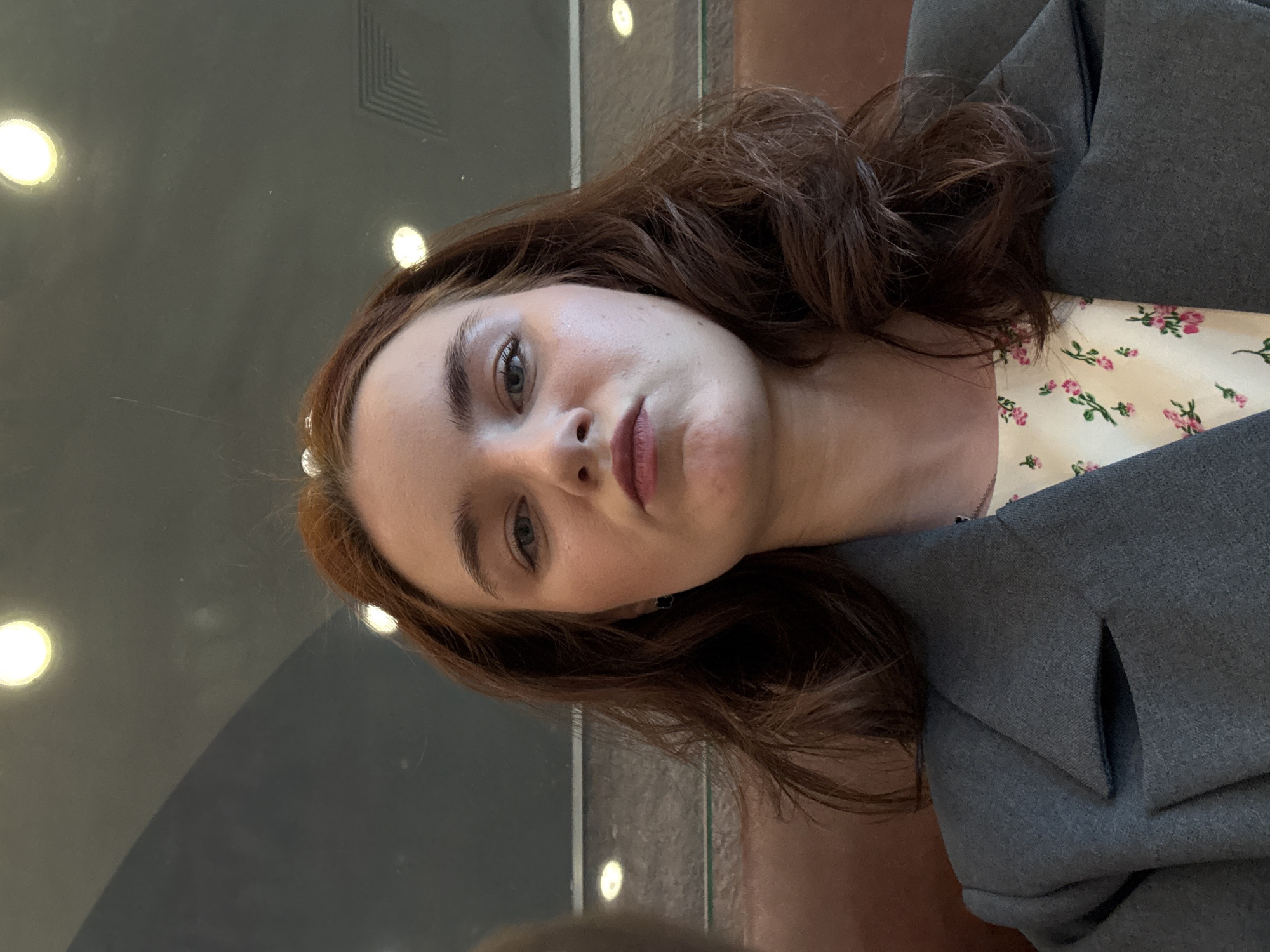 A close-up, horizontally oriented portrait features a young woman with reddish-brown, wavy hair. She is looking directly at the camera with a neutral to slightly serious expression, showcasing her light-colored eyes and well-defined eyebrows. Her head is tilted 90 degrees to the left within the frame.
She is wearing a dark grey blazer over a light-colored top adorned with a subtle floral pattern of small pink blossoms and green leaves. The background is dark and appears to be a reflective surface, possibly glass or polished stone, showing circular reflections of bright lights. In the upper left area of the background, a faint geometric pattern of angled lines is visible. The lighting suggests an indoor setting with artificial illumination. No text is visible in the image.