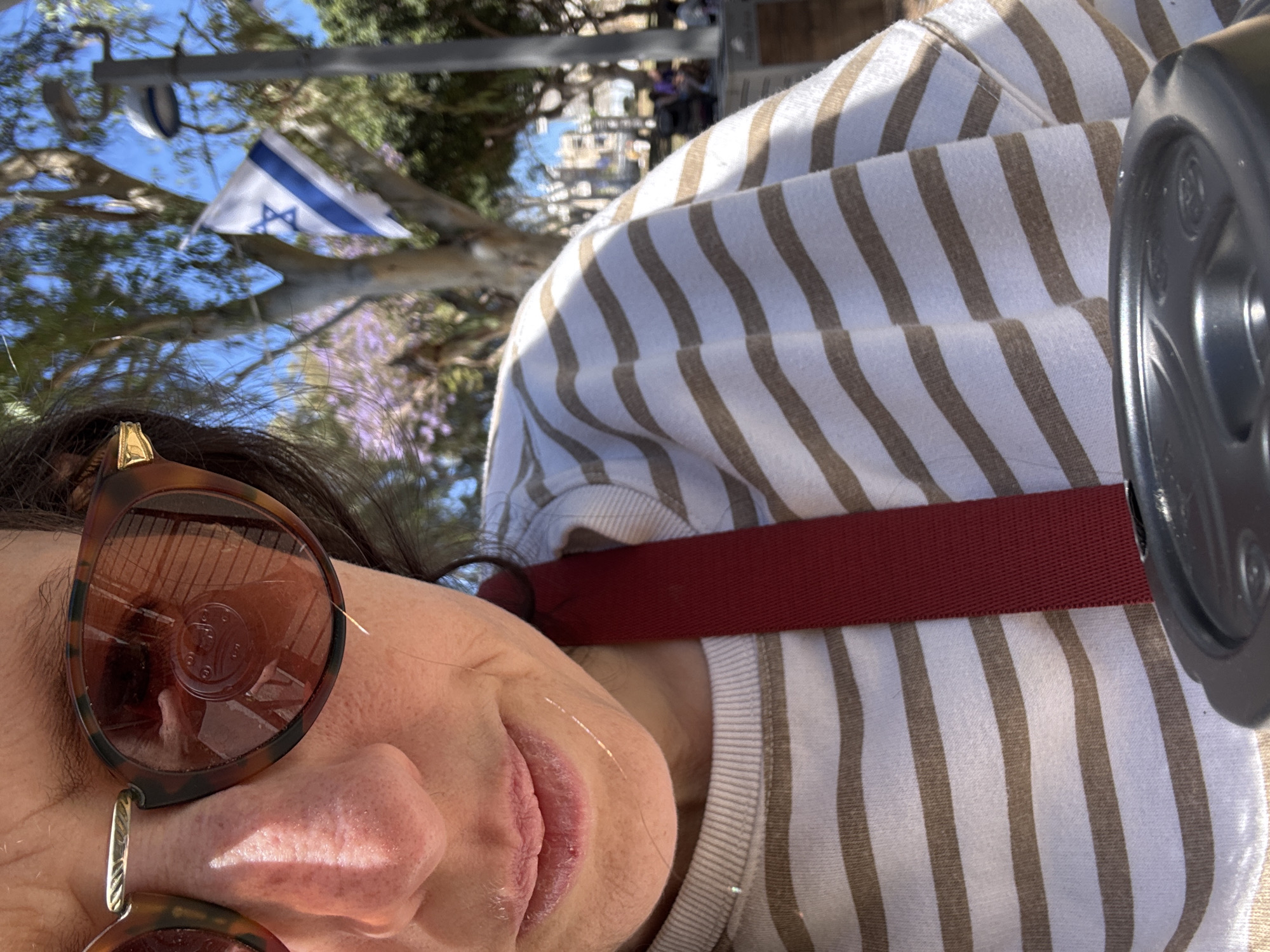 This close-up image, taken in Tel-Aviv, Israel, features a person partially visible outdoors under a bright, clear blue sky. The photograph is oriented vertically, rotated 90 degrees clockwise.
The main subject is a person, likely female, positioned in the foreground. Her face is partly shown, with large, tortoiseshell-patterned sunglasses obscuring her eyes. Her nose and lips are visible, with faint highlights of sunlight on her skin. Dark hair frames her face. She is wearing a light-colored, long-sleeved top with wide, vertical brown stripes. A solid red strap, possibly from a bag or harness, crosses her upper body diagonally over the striped garment. On the far right edge of the frame, a dark gray, circular object, potentially a wheel or part of a device, is partially visible.
The background reveals a sunny outdoor setting with a clear blue sky. Tree branches with green leaves are scattered throughout, and some trees appear to have delicate purple blossoms, possibly Jacaranda. Most notably, a small Israeli flag is seen flying in the background, suspended from a pole. The flag is white with two horizontal blue stripes and a blue Star of David in the center, clearly identifying the location as Israel. The bright sunlight indicates it is daytime. No discernible text is visible in the image. The overall scene suggests a casual moment outdoors on a pleasant day.