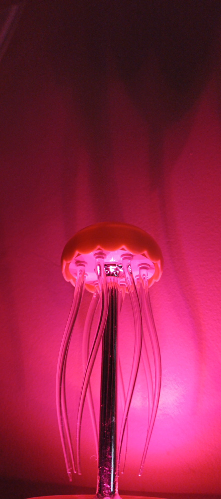 An illuminated jellyfish lamp, colored in shades of pink and red, is positioned against a light pink background. The lamp features a semi-spherical orange 'head' or top, from which several clear, curved strands hang down, resembling tentacles. The head is lit from within, casting a pinkish glow. A vertical chrome or metallic pole supports the head. No people are present in the image. There is no indication that this is located in the city of Chaikovsky, Russia.