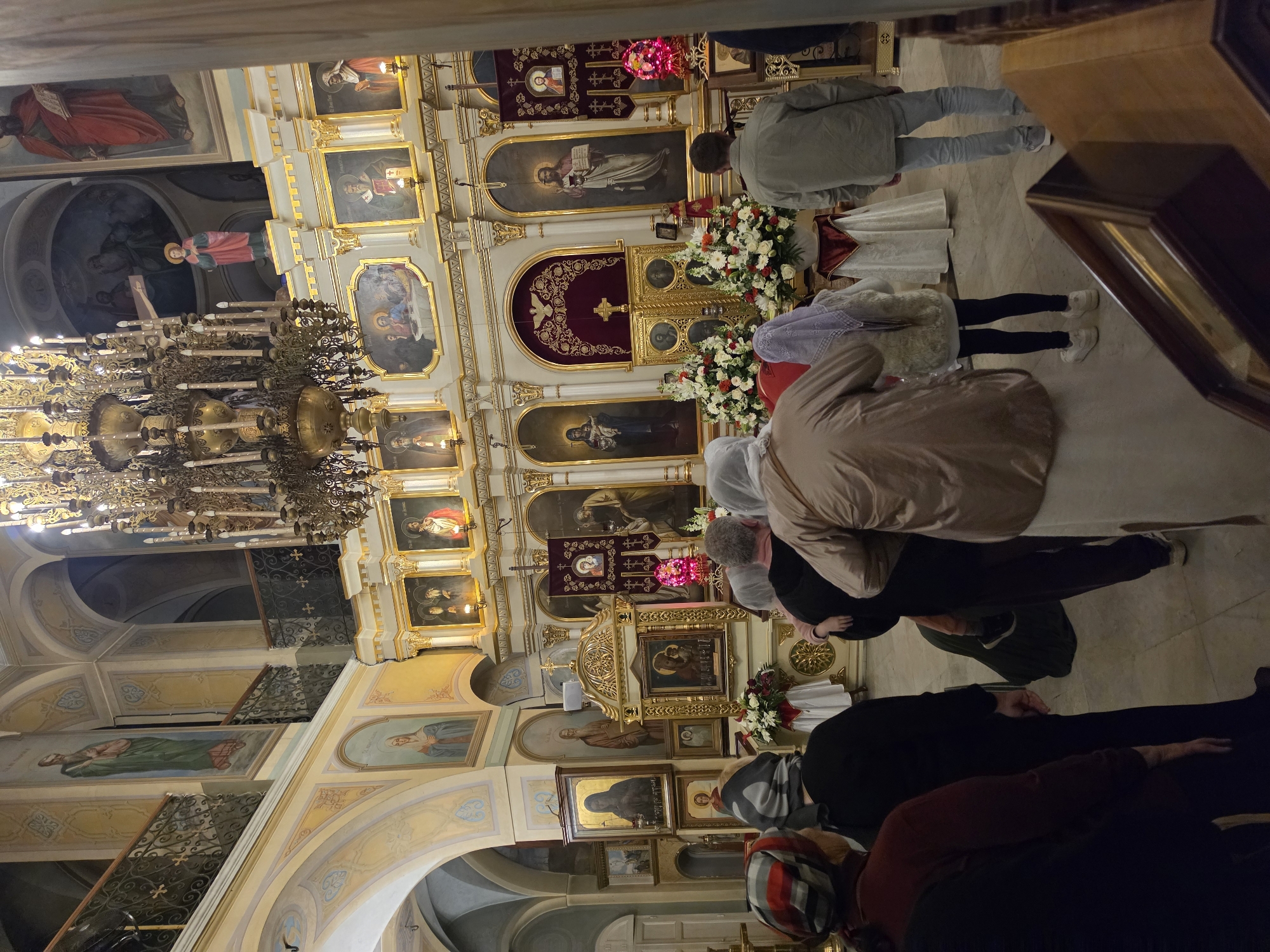 A group of people are inside a decorated Orthodox church. The walls are adorned with religious icons and paintings in golden frames. A large, ornate chandelier hangs from the ceiling. Some people are kneeling or bowing in prayer, while others are standing. Red and white flowers are arranged near the altar.
The location is Tel Aviv, Israel.
