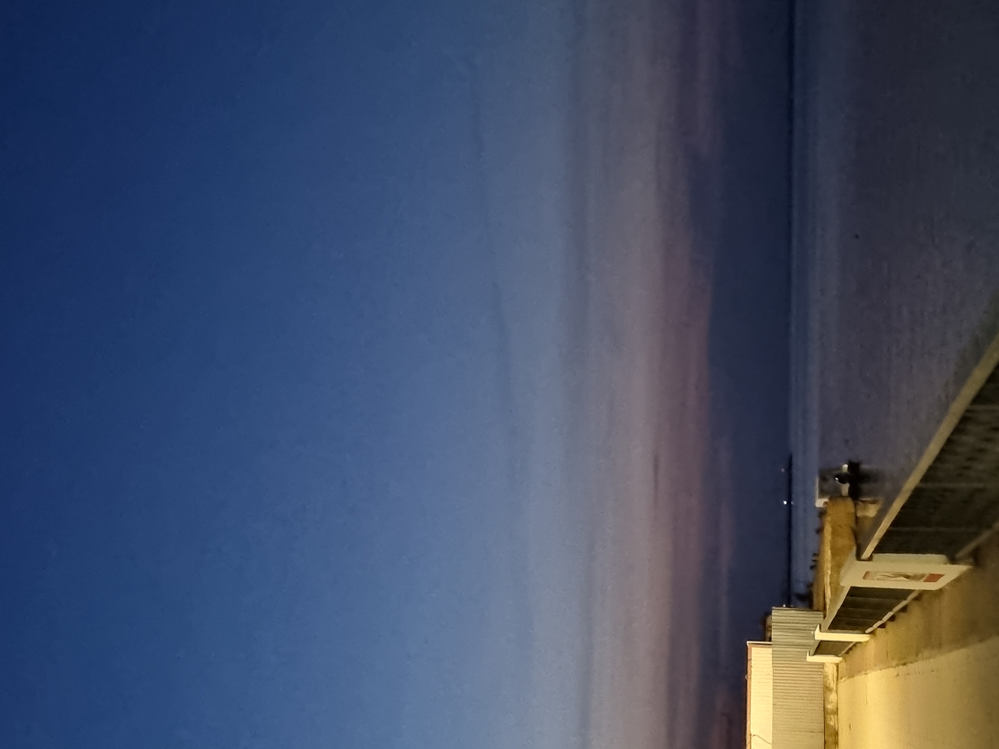 Here is a detailed description of the image:
The image is taken at twilight, with a blue sky transitioning to pink near the horizon. A body of water, likely a river or lake, stretches across the scene. Along the right edge of the image, there is a structure, possibly a building or a dock extending into the water. There are faint lights visible in the distance. Based on the location, this is likely the waterfront area of Chaikovsky, Russia.