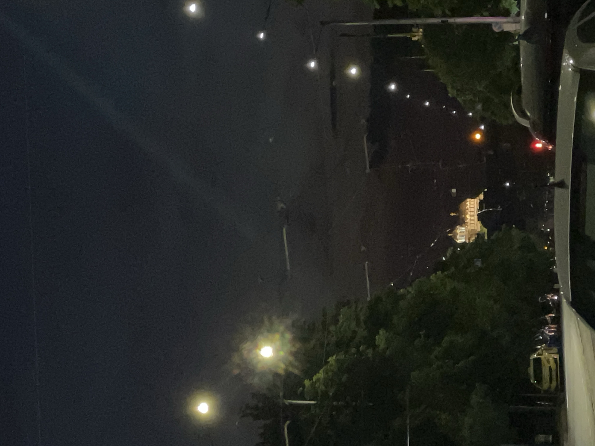 A nighttime image shows a view of a street in Sofia, Bulgaria. A car is parked on the right side of the frame, with the street stretching out into the distance. Streetlights illuminate the road, casting a glow on the surrounding trees. In the distance, a large building is visible, possibly a government building or monument, lit up against the night sky. A crescent moon is visible in the upper left corner of the frame.