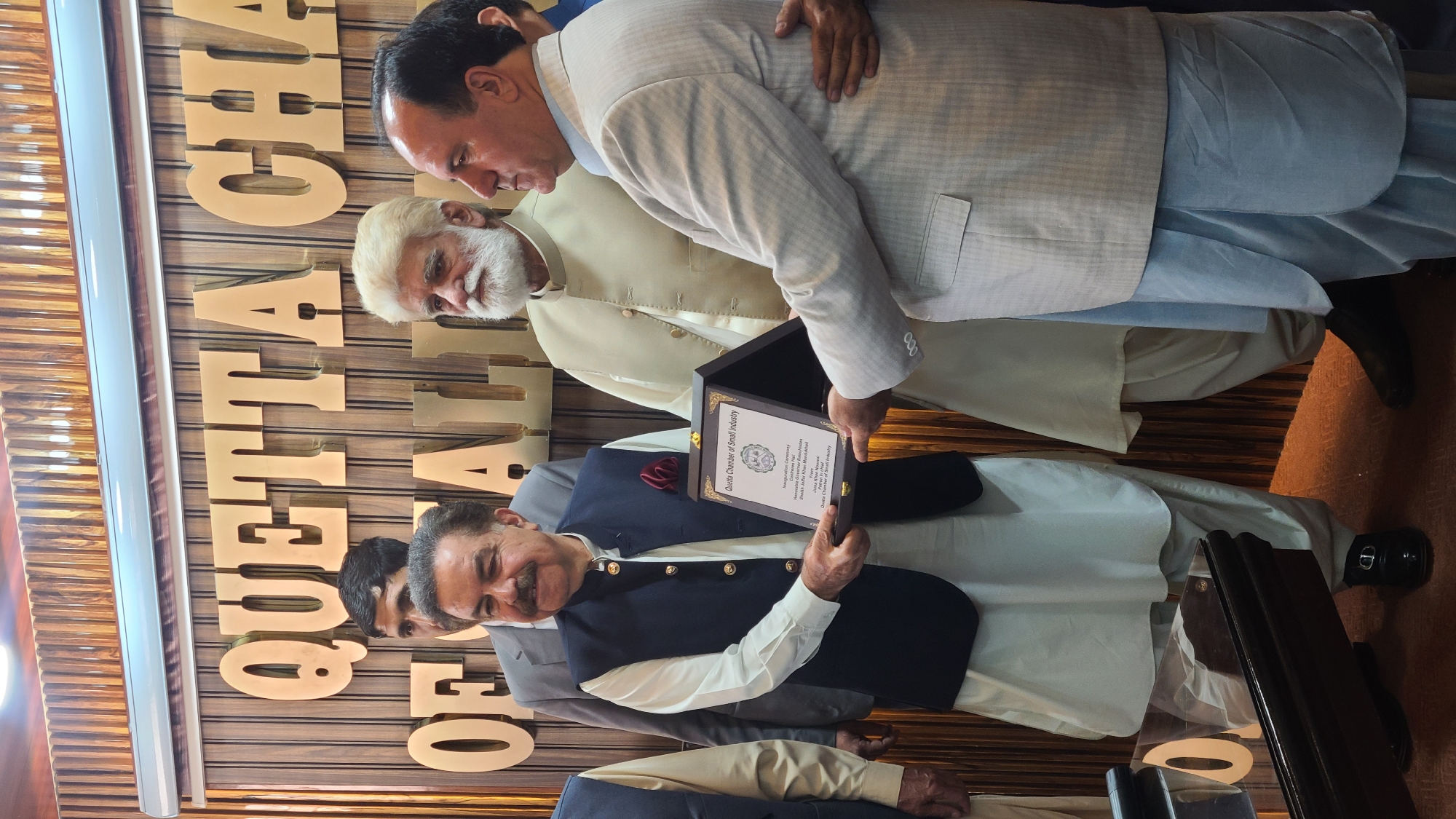 A photograph shows a group of men posing with an award. A man in a dark vest and light-colored traditional Pakistani attire holds an open award box with a certificate inside. The certificate indicates that it is an award from the Quetta Chamber of Small Industry. The city of Quetta City Tehsil, Pakistan, can be inferred from the words "QUETTA CHA" displayed prominently in gold-colored block letters on the wall behind them. Several other men stand behind the man holding the award, dressed in similar traditional Pakistani attire. The room appears to be indoors.
