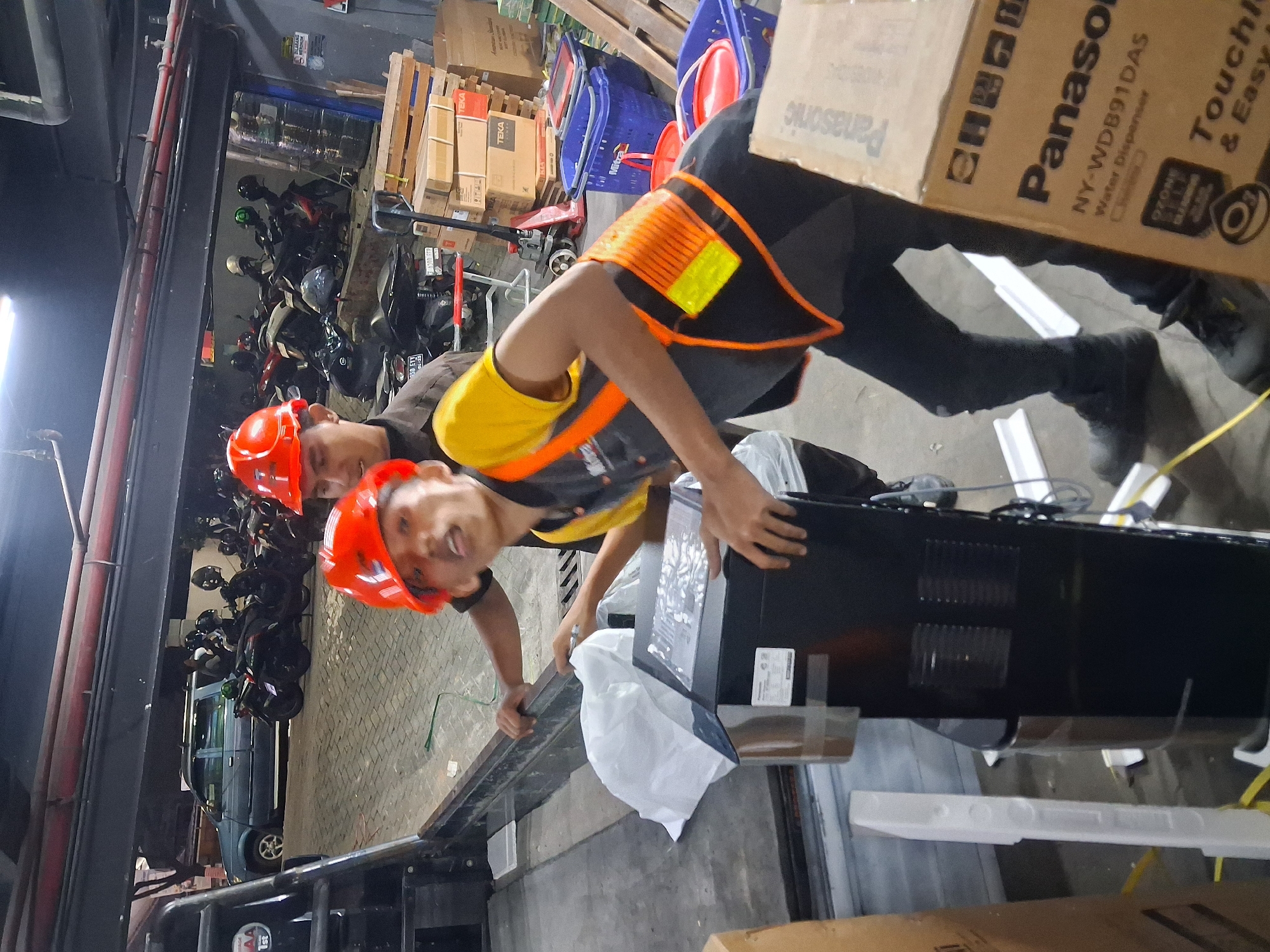 In a covered outdoor area, possibly a loading dock or warehouse entrance in Depok, Indonesia, two young men wearing orange safety helmets and work vests are engaged in moving a water dispenser. The man in the foreground, positioned to the right, is holding the black water dispenser, which has a white plastic wrap partially covering it. He is wearing an orange and black safety vest over a yellow shirt and dark pants. The other man, to the left, is also wearing a safety helmet and is partially visible as he assists with the dispenser. In the background, a dark-colored SUV is parked, and a row of parked motorcycles is visible. Stacks of cardboard boxes, some labeled "TEKA," are present, along with blue and red plastic baskets. The ground is paved with interlocking bricks. The lighting suggests it is either evening or a dimly lit indoor/outdoor space.