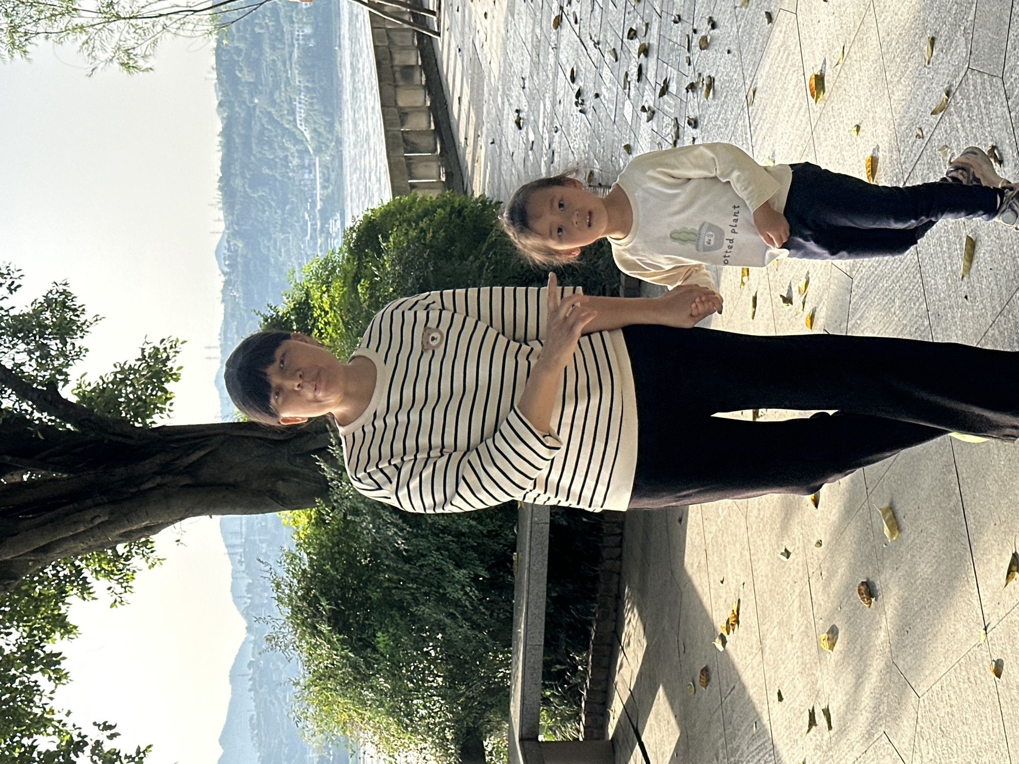 The image depicts an adult and a child standing on a paved surface, likely a walkway. The adult is wearing a horizontally striped shirt and dark pants, and the child is wearing a light-colored long-sleeved shirt with a graphic and dark pants. They are holding hands. Behind them is a view of a river or waterway with tree-covered hills in the background. The location is Jiangjin, China. Fallen leaves are scattered on the pavement.