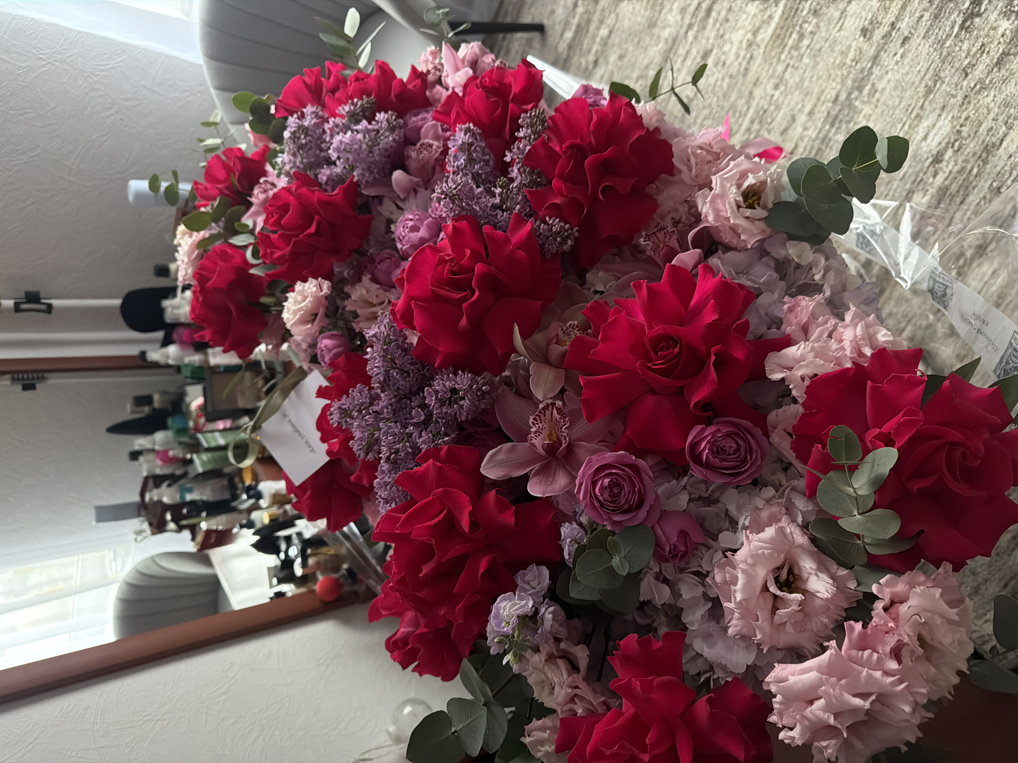 A close-up shot depicts a bouquet of flowers arranged on a surface, likely indoors. The bouquet features a variety of flowers, including red roses, light pink carnations, purple lilacs, and other small purple and white blossoms. Green eucalyptus leaves are interspersed among the flowers. The bouquet has a clear plastic wrapping. In the background, a blurred reflection in a mirror reveals a shelf or table cluttered with various objects. The location cannot be determined as Moscow, Russia based on the provided image alone.