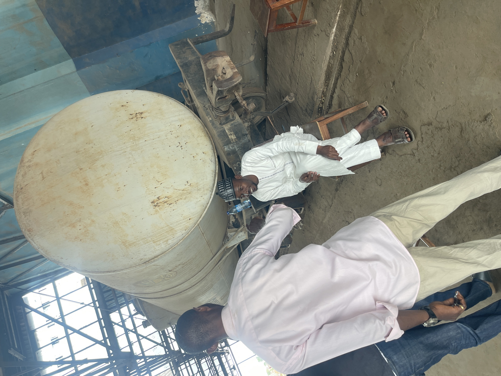 Two men are present in an industrial setting, likely in Zaria, Nigeria. One man, dressed in a white tunic and pants, is reclining on a wooden structure. He has on brown sandals. Beside him is a water bottle. Another man in a pink long-sleeved shirt is standing and holding a small object. A large, cylindrical, possibly metallic object is nearby, possibly a large water tank. A weathered industrial machine is also visible. The scene is indoors, under an industrial structure with exposed metal beams.