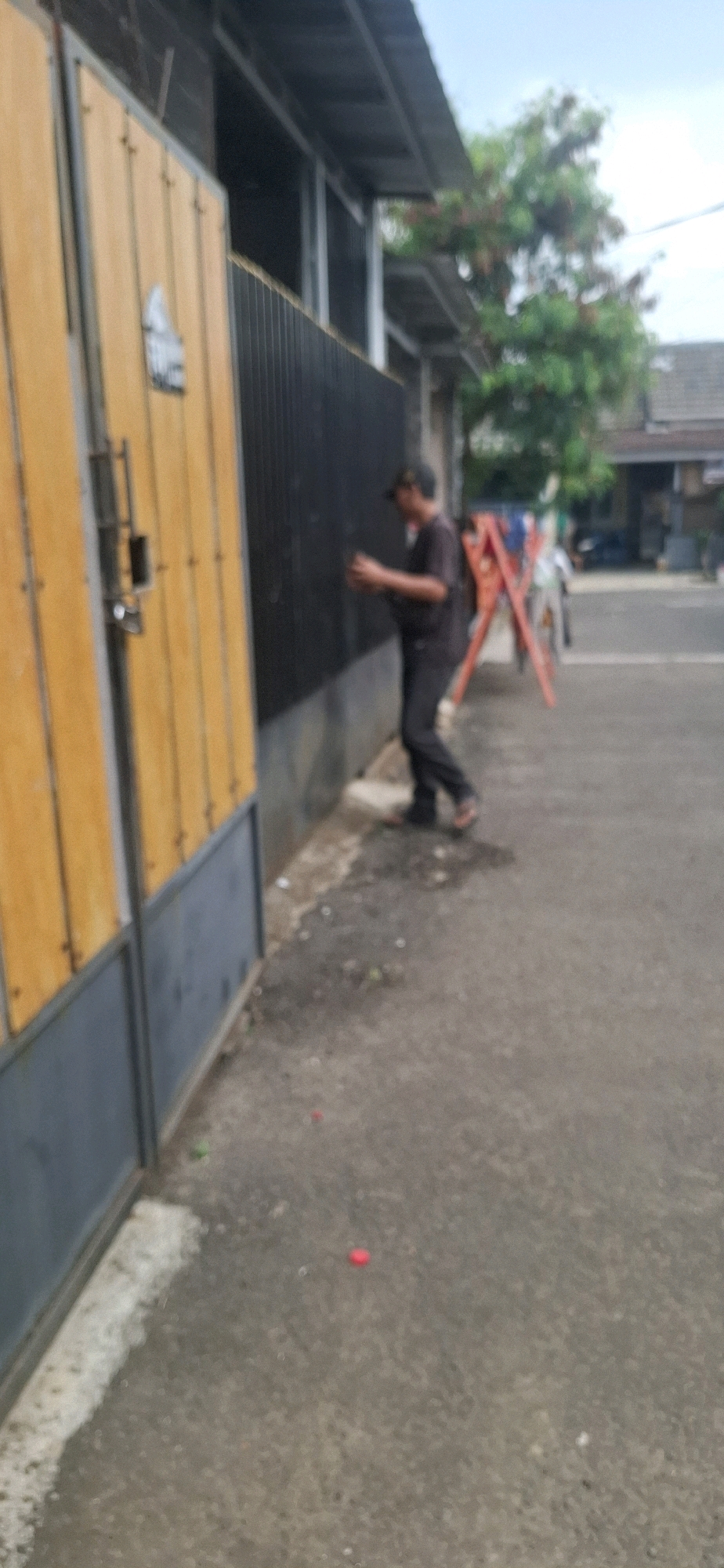 In Cianjur, Indonesia, a person wearing a dark t-shirt, dark pants, and a cap is standing near a black fence. They are holding a phone or similar device and appear to be interacting with the fence. To the left of the fence is a yellow gate with vertical planks and a grey base. In the background, a red A-frame ladder is visible. The scene takes place on an asphalt surface. There are some small red and green objects on the ground, possibly debris. Trees and buildings are visible in the distance. The sky is cloudy.