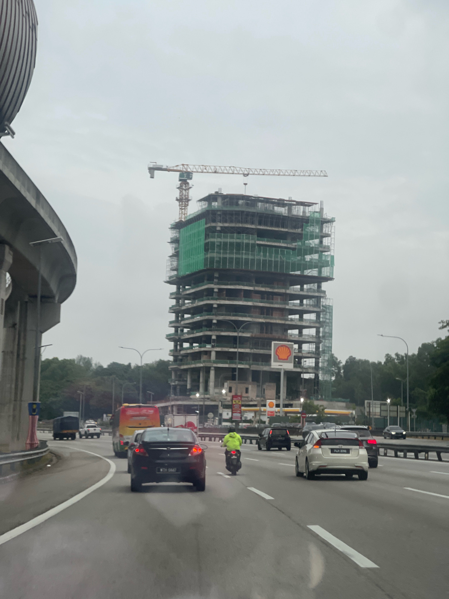 The image captures a view from a highway in Shah Alam, Malaysia, with a multi-story building under construction dominating the skyline. A large yellow and orange construction crane is positioned above the building, with its long arm extended horizontally. The building itself is a skeletal structure of concrete and steel, with green netting draped across some of its levels, suggesting ongoing construction work. A prominent Shell logo is visible on the side of the building, indicating a commercial establishment is planned.
The highway is busy with traffic. Several cars are driving on the road, including a black sedan in the foreground with license plate "WTH 9447". A white sedan with license plate "PLH 3018" is in the middle lane. A motorcycle with a rider wearing a bright green jacket is also on the road. Further back, a yellow and red bus is visible, along with other vehicles. The sky is overcast, contributing to a subdued and hazy atmosphere. Trees line the sides of the highway, and an elevated concrete structure, possibly part of a flyover or overpass, curves into the frame from the left. The overall scene depicts a typical urban environment during daylight hours, characterized by infrastructure development and vehicular movement.