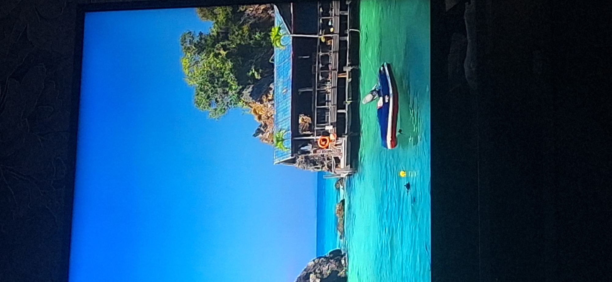 The image depicts a vibrant tropical scene with a clear blue sky and turquoise water. In the foreground, a jet ski with red and white accents is floating on the water. To the left, two dark figures, possibly swimmers or divers, are visible near a yellow float. In the mid-ground, a weathered wooden stilt house with a blue roof is situated on a rocky outcrop, adorned with a life preserver. Two small palm trees flank the structure. The background features lush green trees and rugged brown rocks, suggesting a coastal or island environment. The image does not contain any recognizable landmarks or features specific to Bila Tserkva, Ukraine.