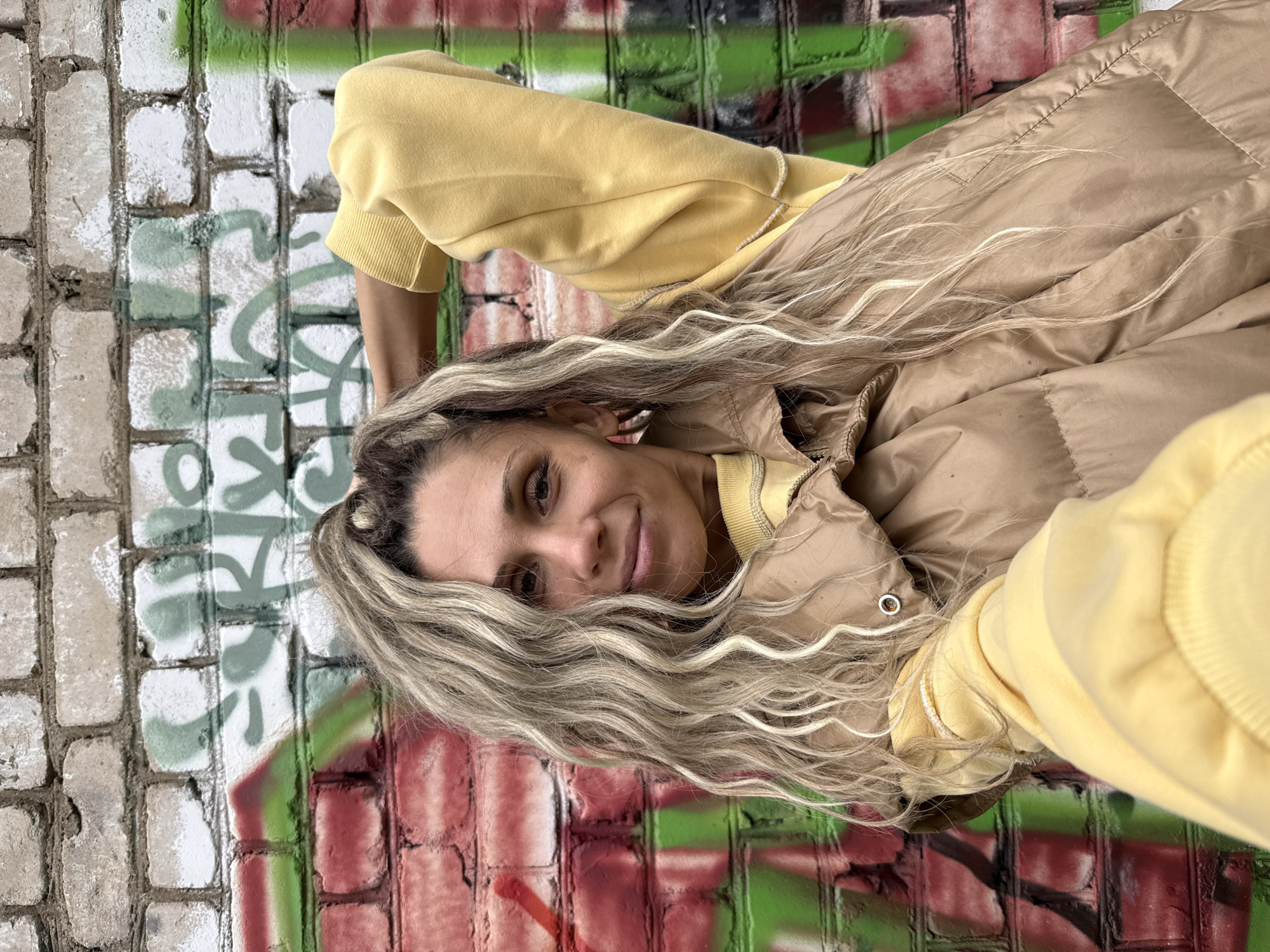 A medium shot captures a woman with long, wavy blonde hair standing in front of a graffiti-covered brick wall in Rusanovo, Russia. She is wearing a yellow sweatshirt and a tan puffer vest. Her left arm is raised behind her head, and she is looking towards the camera with a slight smile. The brick wall behind her is painted with various colors of graffiti, including red, green, and white. The lighting suggests it is daytime.