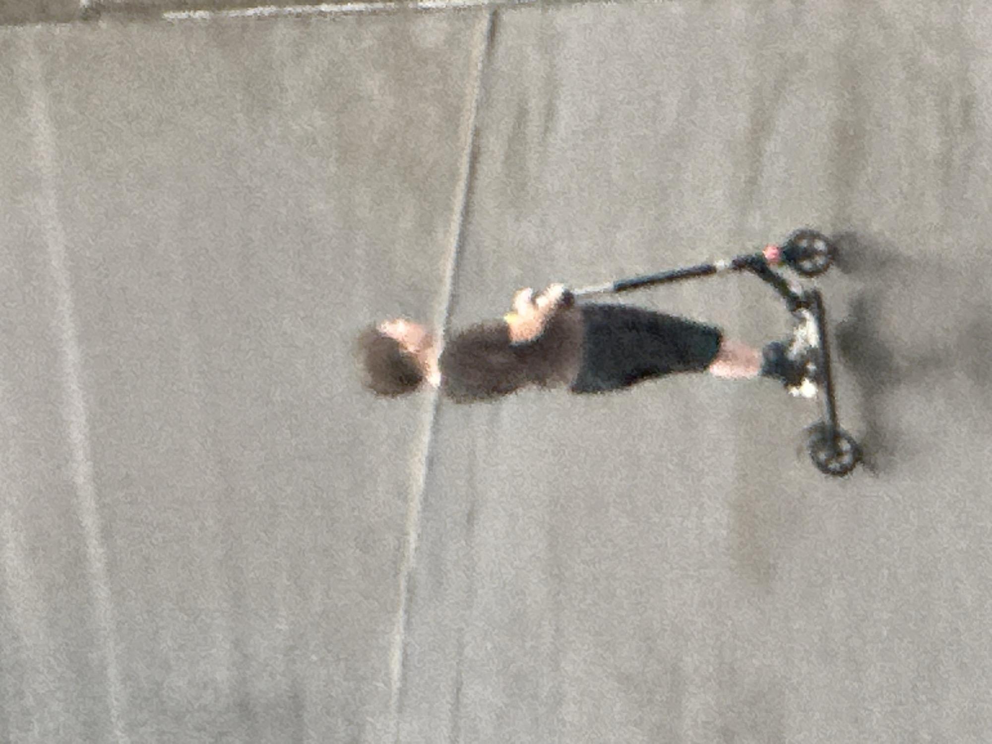 A young person wearing a dark t-shirt and dark shorts stands on a scooter on a paved surface. The scooter has a dark frame and two wheels, one visible at the front and one at the rear. The person's face is not clearly visible, but their hair appears dark and curly. The background is a textured, gray paved area with a faint horizontal line visible in the distance. This scene is taking place on a paved outdoor area, possibly a street or plaza, in the city of Podolsk, Russia.