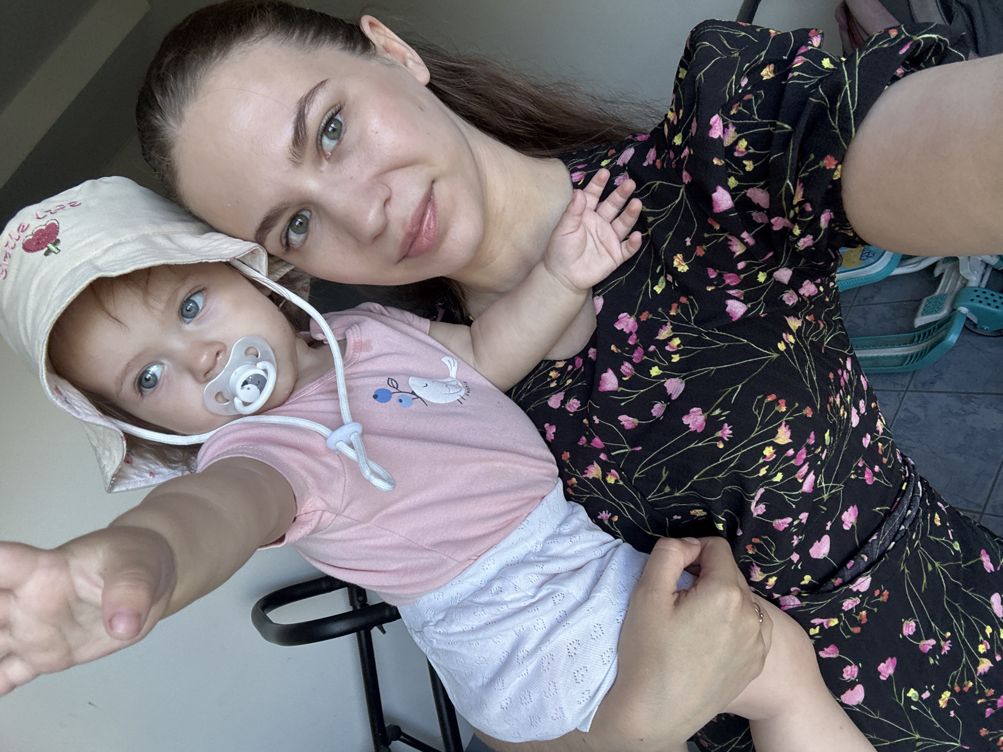 A woman is holding a baby in an outdoor setting. The woman is on the right side of the frame, looking towards the camera with a neutral expression. She has brown hair pulled back and is wearing a black dress with a floral print. The baby is on the left side of the frame, facing away from the camera and reaching upwards. The baby is wearing a light-colored sun hat and a pink onesie with white lace shorts. A pacifier is in the baby's mouth. The background is out of focus but appears to be a wall and some furniture. This scene is unlikely to be in Chaikovsky, Russia, as the image depicts an indoor setting, not an outdoor city location.