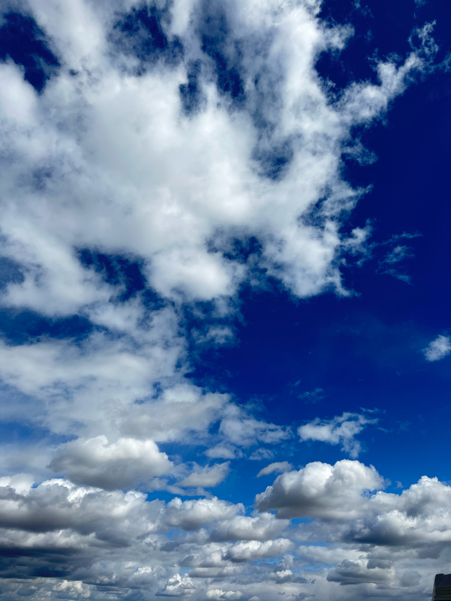 The image depicts a sky filled with fluffy white and grey clouds against a deep blue sky. The clouds are varied in size and density, with some appearing wispy and others more solid and voluminous. The lighting suggests it is daytime, with sunlight illuminating the clouds from above, creating shadows and highlights. The scene appears to be an outdoor shot looking upwards, capturing a dynamic cloud formation. There are no people, animals, or man-made structures clearly visible, though the bottom edge of the image hints at a possible rooftop or elevated surface. The location of Цабелевка, Kazakhstan, provides a geographical context for this atmospheric scene.
