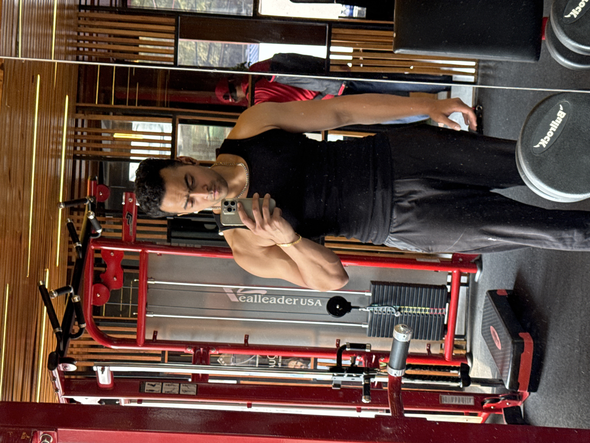 A man is reflected in a mirror in a gym. He wears a black tank top and dark pants. He holds a smartphone, presumably taking a picture. The background of the reflection reveals gym equipment, including a red weight machine labeled "RealLeader USA," dumbbells labeled "Bullrock," and wooden wall paneling. Another individual, wearing a red cap and shirt, is also visible in the reflection. The location cannot be determined as Kolkata, India based on the given image.